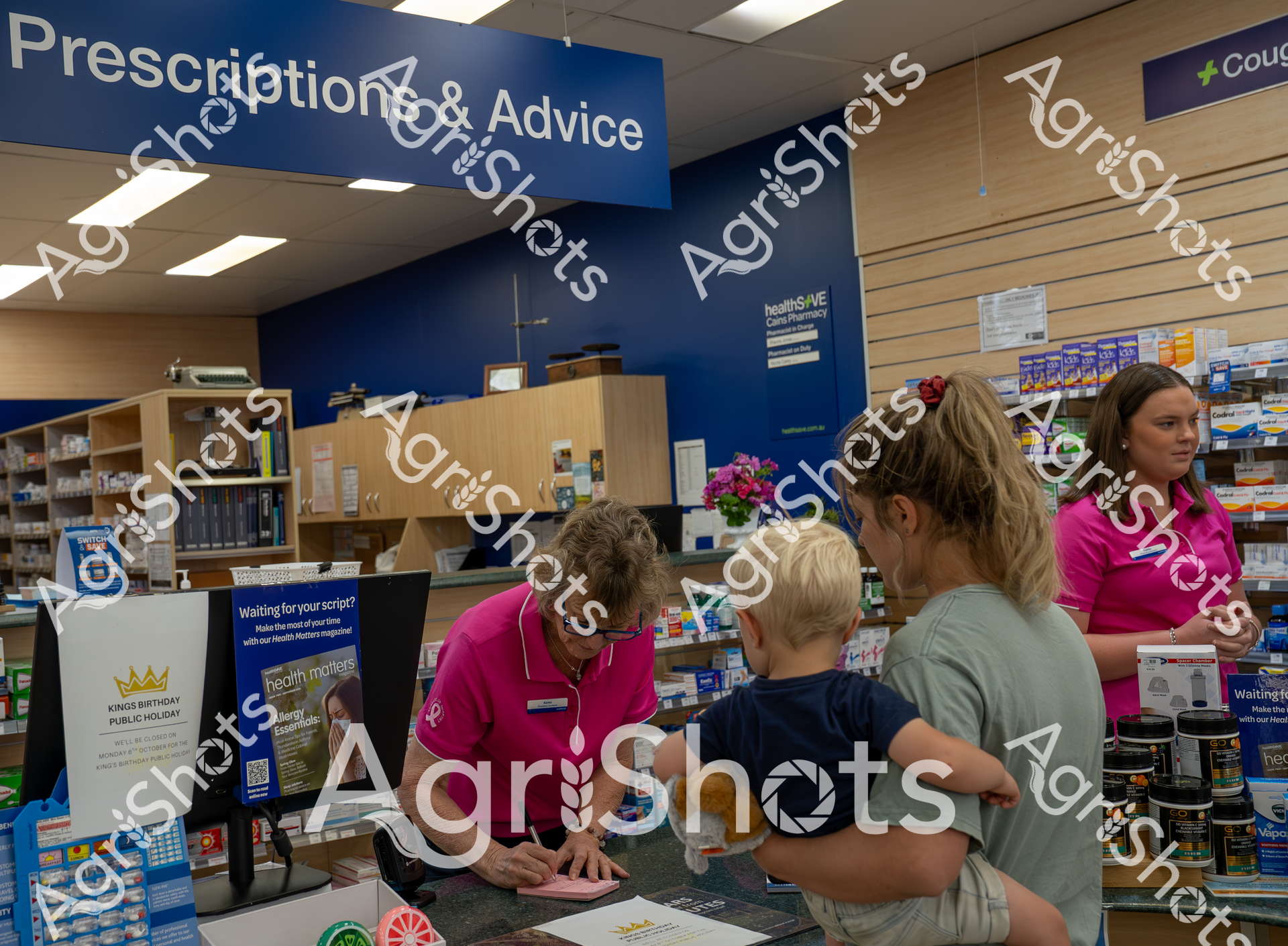 A pharmacy scene showing a woman in a pink shirt assisting a customer with a child at the prescription counter. Shelves of medication and health products line the background, with a "Prescriptions & Advice" sign overhead. The pharmacy appears well-organized and professional, with staff ready to help customers.