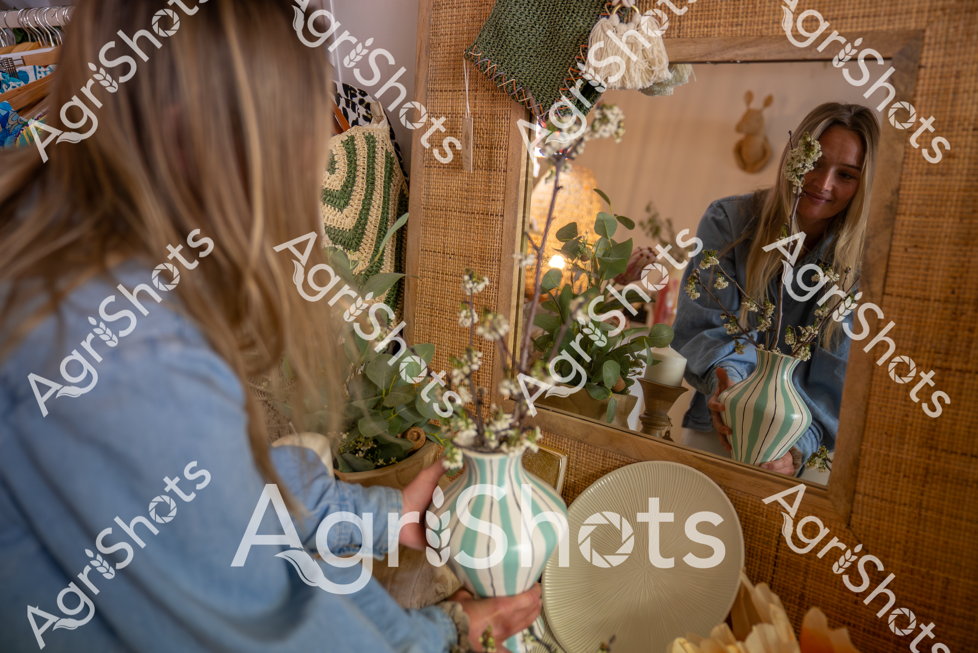 A woman in a blue denim shirt arranges delicate white flowers and green foliage in a striped turquoise and white vase. She is standing near a woven backdrop and a mirror, creating a soft, natural aesthetic with warm lighting and botanical elements. Her reflection shows her carefully positioning the floral arrangement with a gentle, focused expression.