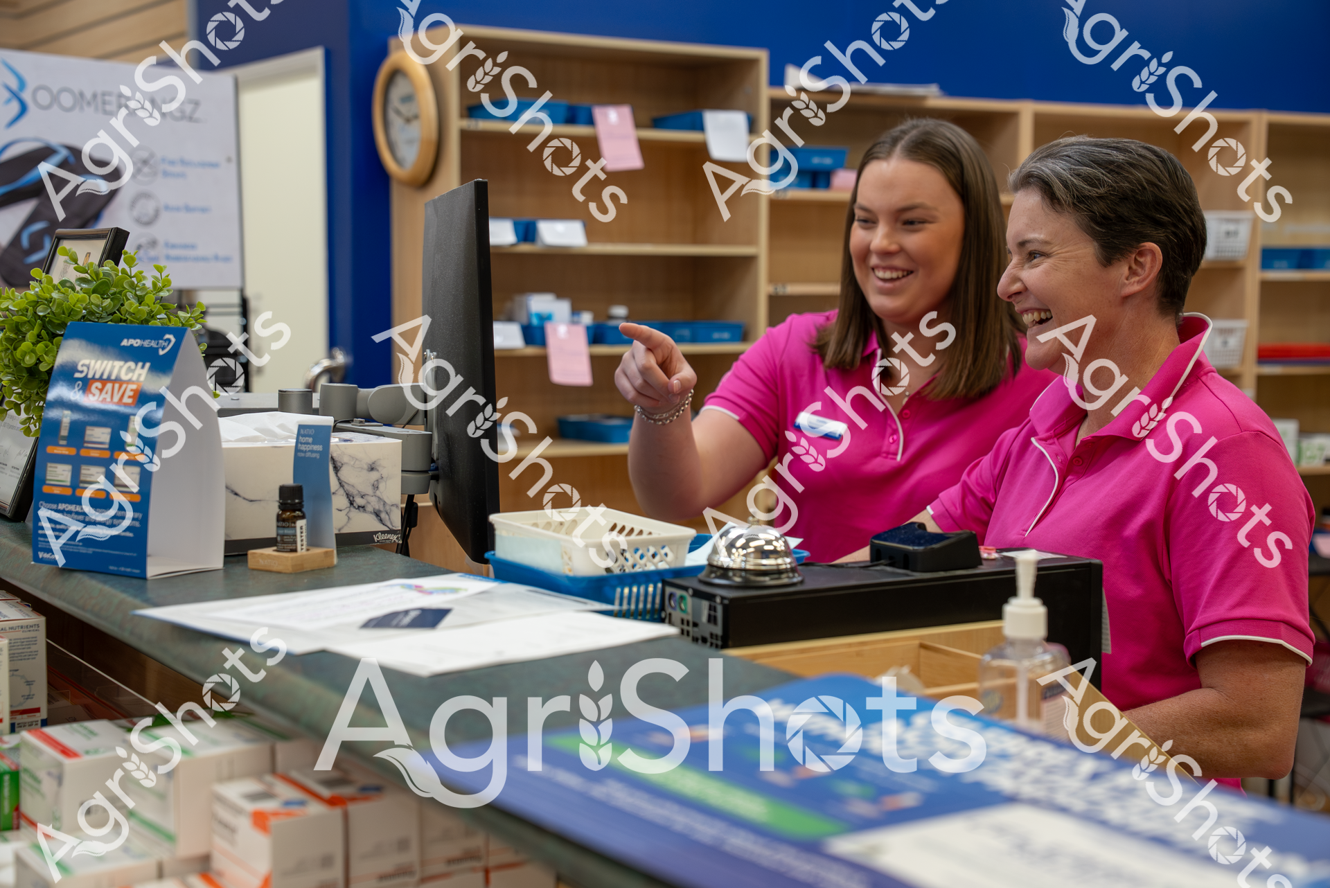 Two women in matching bright pink shirts work together at a retail counter, laughing and pointing at something on a computer screen. The scene appears to be in a store with shelves and product displays, suggesting they are coworkers enjoying a moment of camaraderie during their workday.