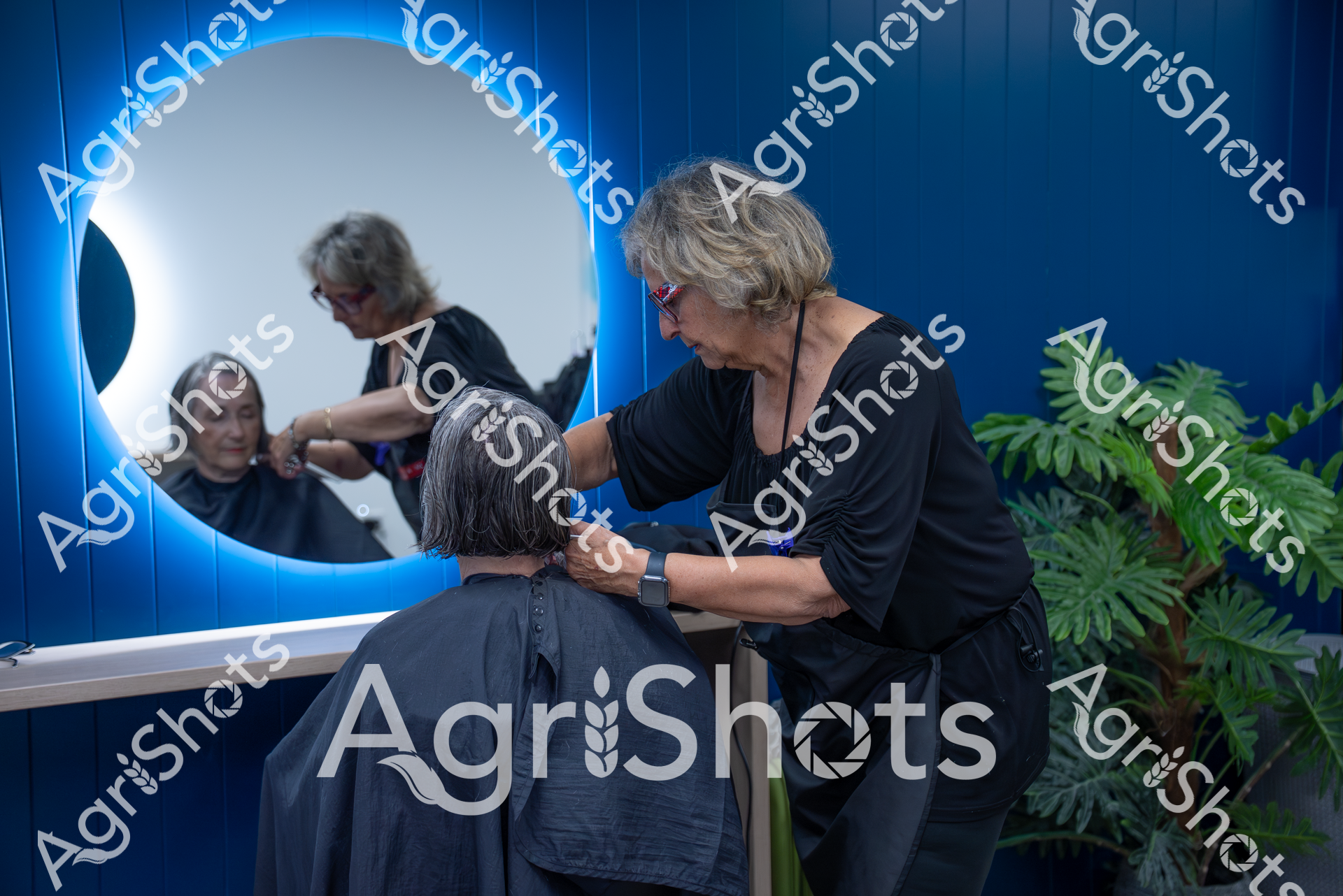 A professional hairstylist works on a client's hair in a modern salon setting. The stylist, wearing black, carefully cuts or styles the client's hair in front of a large illuminated circular mirror with blue ambient lighting. Green plants in the background add a touch of natural elegance to the contemporary salon space.