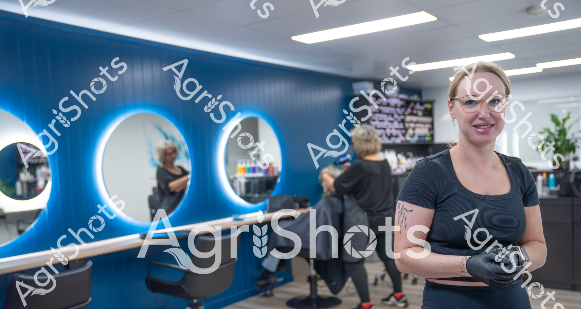 A smiling woman in a black shirt and gloves stands in a modern hair salon with blue-accented walls and illuminated mirrors. In the background, another stylist works with a client, creating a professional and vibrant atmosphere typical of a contemporary hair and beauty workspace.