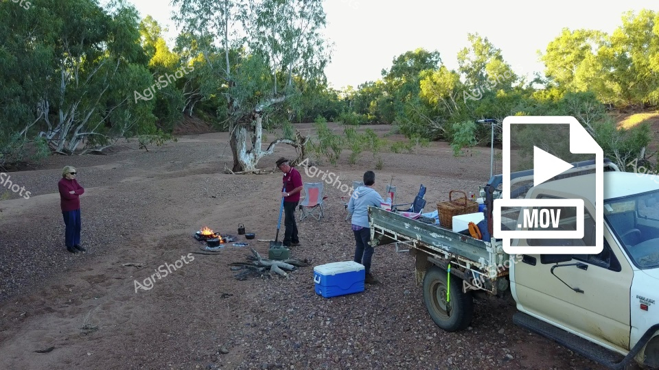 People gather around a campfire at a Queensland station, with camp oven cooking and camping equipment scattered across the dusty ground. A white ute is parked nearby with gear loaded in the back, while men and women station hands prepare for an outdoor meal in the bush setting. The scene captures the essence of remote station life, with trees providing shade and a blue cooler positioned near the cooking area.