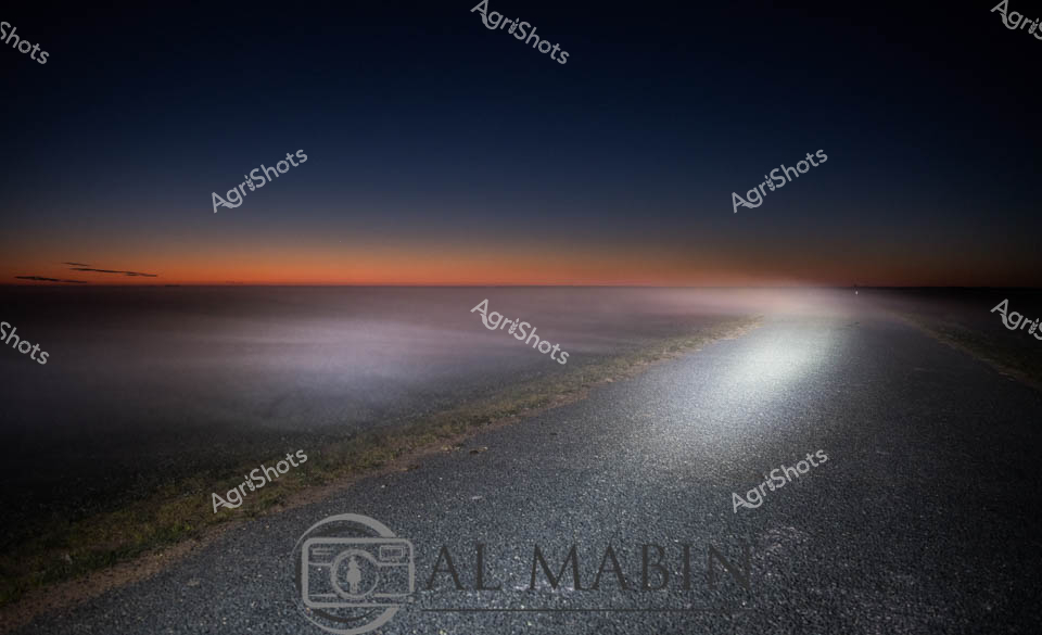 A twilight landscape emerges, revealing a lonely rural road stretching into misty horizons under a gradient sunset sky. The asphalt road cuts through a dark landscape, with a brilliant white light beam illuminating one side, creating a dramatic contrast between darkness and brilliant luminescence. The scene captures the ethereal transition between day and night, with deep blue-black tones gradually blending into warm orange sunset hues near the horizon. Soft fog or mist seems to blur the boundaries between land and sky, lending a mysterious and contemplative atmosphere to the scene. The road appears isolated and expansive, suggesting a remote agricultural or rural setting typical of Australian countryside landscapes. The long exposure and carefully balanced lighting create a sense of stillness and infinite possibility, transforming an ordinary roadway into a poetic visual narrative of transition and silence.