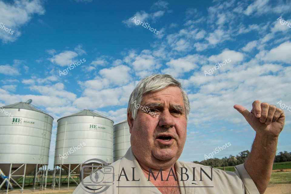 An experienced farmer stands resolute against a brilliant blue sky mottled with scattered white clouds, surveying his agricultural domain. Gleaming HE-branded grain silos rise behind him, their metallic surfaces reflecting the bright midday sunlight of a classic Australian rural landscape. The seasoned agricultural worker gestures with his thumb, pointing toward the storage infrastructure that represents the backbone of his farming operation. His weathered face and crisp white shirt tell a story of decades of agricultural expertise, while the expansive paddocks and precise grain storage systems speak to modern farming precision. The composition captures the intersection of traditional agricultural knowledge and contemporary farming technology, with the silver silos symbolizing agricultural efficiency and economic potential. This portrait embodies the resilience, pride, and technological sophistication of contemporary Australian agricultural enterprise, set against the quintessential wide-open rural landscape of New South Wales.