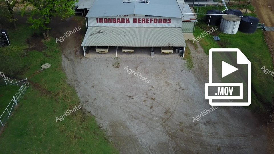 An aerial view of the Ironbark Herefords farm facility, featuring a corrugated iron shed with the farm name prominently displayed. The image shows a graveled yard area surrounded by green grass, with water tanks and fencing visible in the periphery. This overhead perspective provides a comprehensive look at the agricultural property's infrastructure and layout.