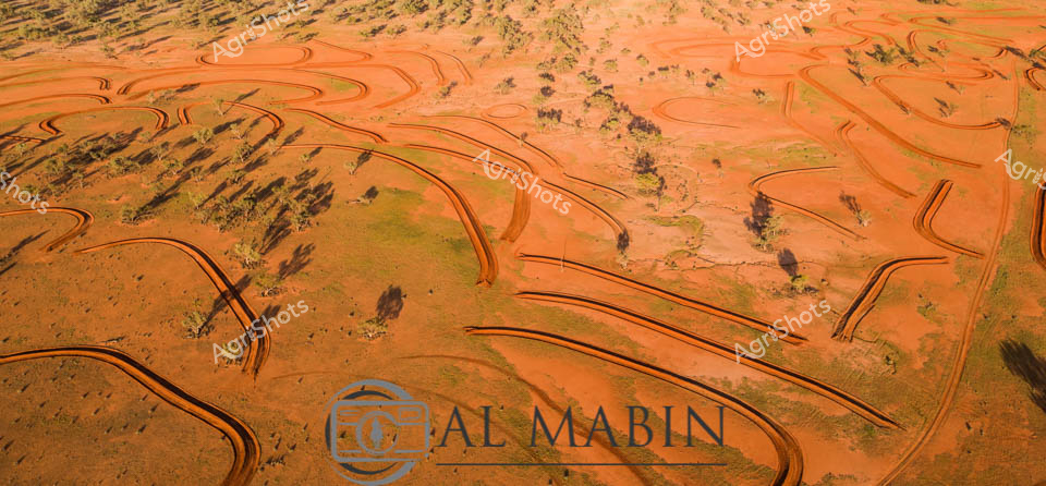 Aerial view of meticulously sculpted contour banks sweeping across the rich terracotta landscape of rural New South Wales, revealing an extraordinary agricultural intervention against the arid inland terrain. Sinuous orange-red earthen curves trace intricate lines across the sparse grassland, creating a mesmerizing pattern of agricultural engineering that follows the natural topography. These carefully constructed erosion control banks wind through scattered native vegetation, transforming the harsh desert environment into a carefully managed agricultural canvas. The undulating earthen lines demonstrate sophisticated land management techniques, protecting precious soil from water erosion and capturing critical moisture in this challenging agricultural region. Vibrant ochre tones dominate the landscape, punctuated by occasional patches of muted green scrub, highlighting the delicate balance between human agricultural intervention and the raw Australian outback environment.