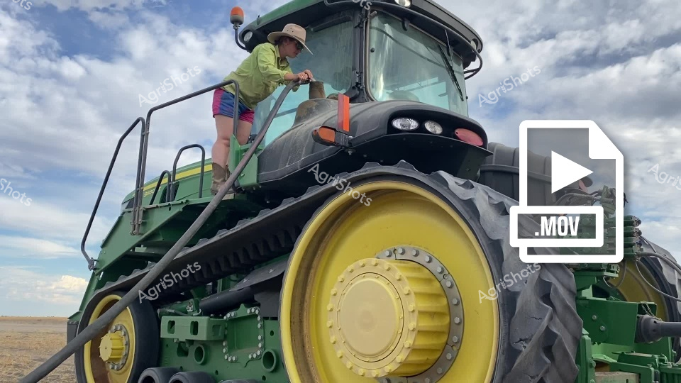 A farm worker in high-visibility safety gear climbs aboard a massive green John Deere tracked tractor in a New South Wales wheat field. The powerful agricultural machine, equipped with distinctive yellow wheel rims and robust track systems, stands ready for grain harvesting operations across the expansive cropped landscape. Cloudy skies stretch overhead as the worker prepares the heavy-duty equipment for another day of wheat harvest, showcasing the scale and precision of modern Australian cropping systems.