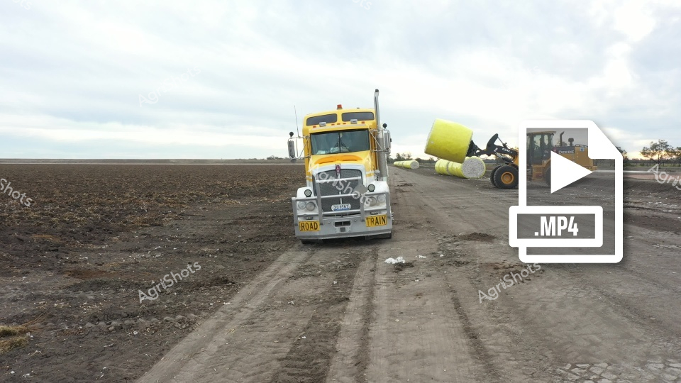 A bright yellow road train truck hauls cotton bales across a dusty agricultural field in New South Wales, with a John Deere loader in the background preparing additional bales. The expansive, flat landscape of Walgett Shire showcases the important cotton harvest season, highlighting the region's agricultural productivity and transportation infrastructure.