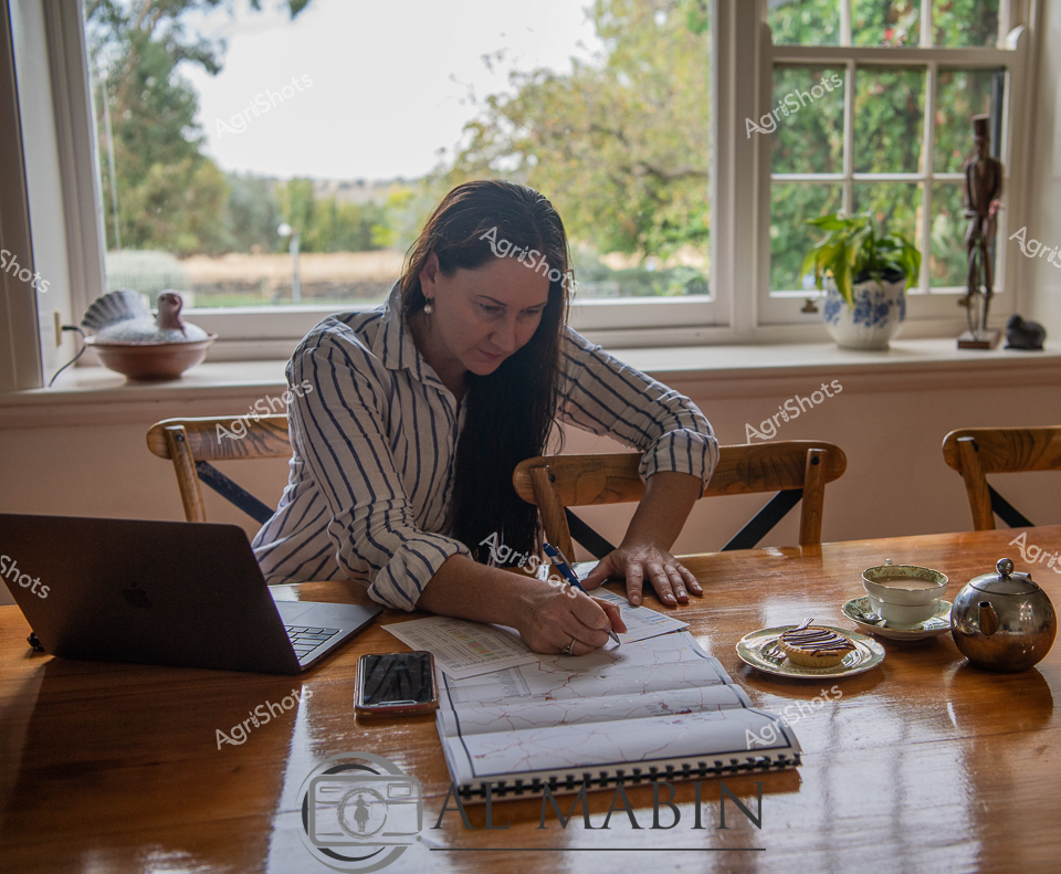 Rural productivity pulses through this intimate workspace, where a focused agricultural professional crafts her day's strategy. Bathed in soft natural light from expansive windows overlooking verdant rural landscape, the scene captures a contemporary farmer or agricultural manager immersed in detailed planning. Her crisp blue-and-white striped shirt speaks to both professional precision and practical rural style, while her careful annotations across extensive documents suggest meticulous enterprise management. A gleaming laptop, smartphone, and an elegant tea service complement her workspace - a sophisticated blend of traditional agricultural documentation and modern digital technology. The warm wooden table, classic ceramic teacup, and glimpse of pastoral scenery through mullioned windows evoke a sense of connection between strategic agricultural work and the living landscape that sustains it. This image celebrates the intellectual labor and quiet determination of women driving agricultural innovation and management in regional Australia.