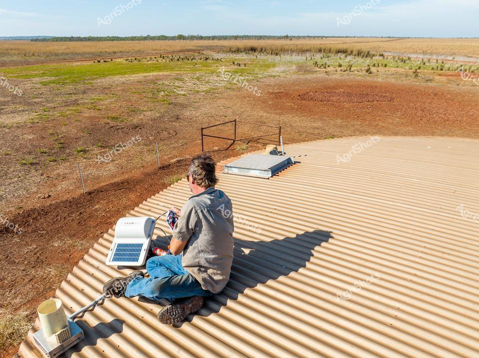 Rural Solar Monitoring in the Top End

A station worker sits cross-legged on a corrugated metal water tank roof, surveying the expansive Northern Territory landscape. Wearing a grey work shirt and blue jeans, the person carefully manages solar monitoring equipment, positioning a small solar panel to capture precise environmental data. The surrounding terrain stretches into a vast, ochre-hued pastoral landscape, with sparse grasslands and scattered vegetation marking the distant horizon. Windswept and sun-bleached, the environment speaks to the challenging conditions of remote Australian agricultural operations. A metal framework surrounds the water tank, providing structural support and a practical mounting point for technical infrastructure. The scene captures the intersection of traditional rural infrastructure and modern renewable energy technologies, highlighting how innovative solutions are being integrated into Australia's agricultural heartlands.