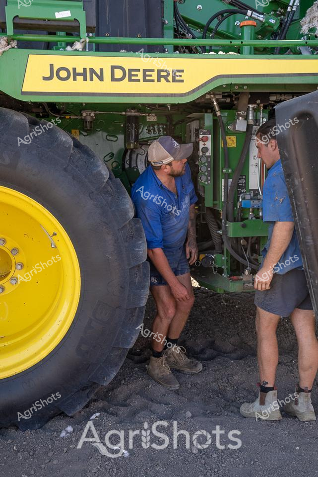 Two agricultural workers stand intently focused between the massive yellow wheel and green framework of a John Deere cotton picker. Dressed in matching blue work shirts and casual shorts, they collaborate closely within the complex machinery, suggesting a coordinated team approach to harvesting. The heavy-duty agricultural equipment dominates the frame, its industrial green and yellow color palette instantly recognizable as John Deere's signature branding. Oversized black rubber tires and intricate hydraulic systems highlight the sophisticated engineering required in modern cotton harvesting technology. Dusty ground beneath their work boots indicates a rural setting, likely in one of NSW's prominent cotton-growing regions like Walgett Shire. The scene captures the precision and technical expertise demanded in contemporary agricultural operations, where human skill and advanced machinery work in seamless partnership to efficiently process critical agricultural crops.