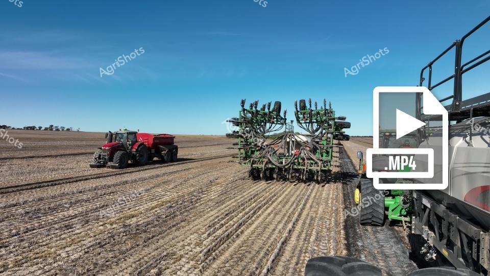 Agricultural seeding operation in Western Australia's golden cropping season, featuring a red tractor pulling a massive green John Deere air cart and seeding implement across a prepared field under a clear blue sky. The precision farming equipment moves methodically, demonstrating modern agricultural techniques in a vast, flat agricultural landscape. Advanced machinery works to plant the next crop with efficiency and technological sophistication.
