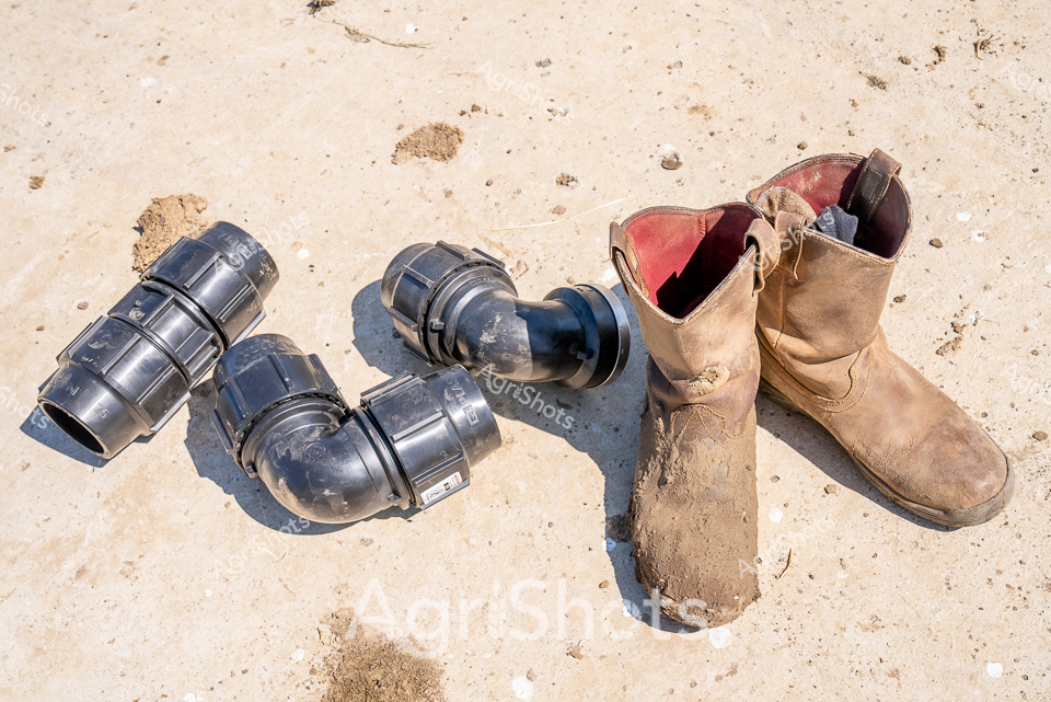 Sun-baked earth cradles a collection of robust Plasson pipe fittings beside a weathered work boot, capturing the stark beauty of remote Western Australian agricultural infrastructure. Navy blue compression joints curve and connect with precise engineering, their industrial form a stark contrast to the soft sandy ground. A well-worn leather boot, faded to a dusty tan from countless days of hard labor, anchors the scene with its rugged authenticity. These hydraulic and irrigation components speak to the sophisticated water management systems essential in Australia's challenging agricultural landscapes, where every drop of water is precious. The metallic fittings' smooth, curved surfaces reflect the harsh midday light, while the boot's scuffed leather tells stories of endless kilometers walked across station country. This image encapsulates the pragmatic resilience of agricultural workers who transform unforgiving terrain through careful engineering and persistent effort.
