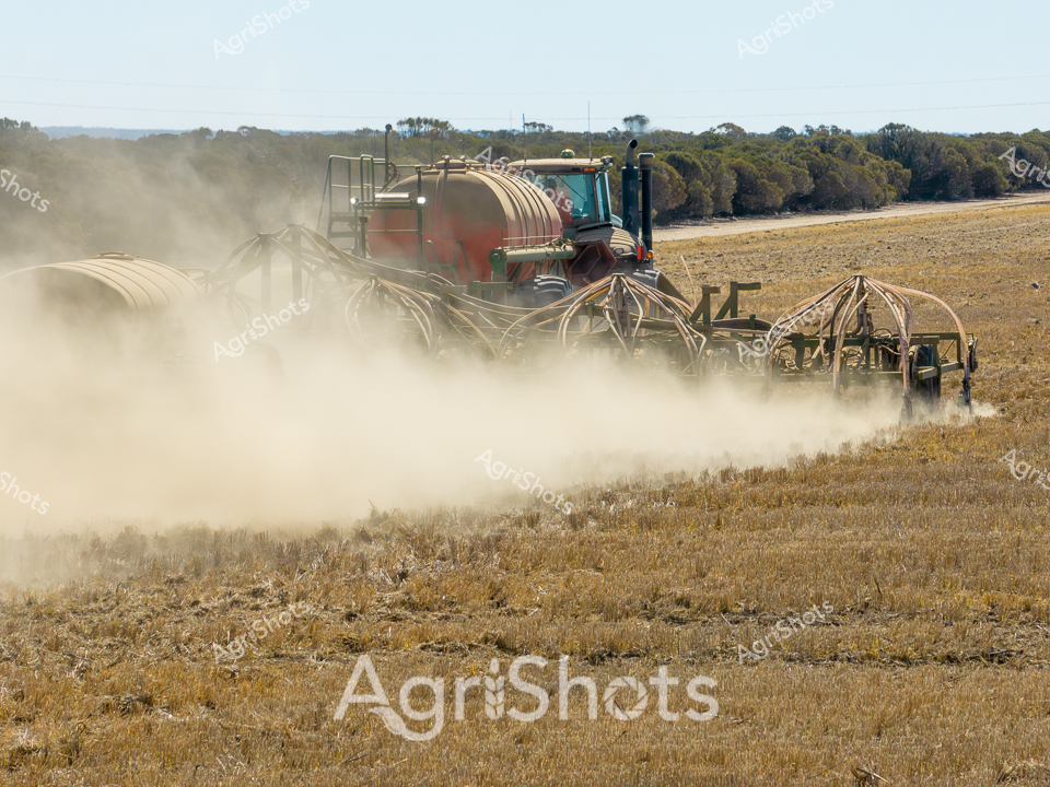 Billowing clouds of dust rise from the dry paddock as a robust John Deere tractor glides across the golden stubble-covered landscape of Western Australia. The agricultural air cart, mounted with large cylindrical tanks, trails behind, dispersing seeds with precision across the sun-baked earth. Sweeping lines of trees mark the distant horizon, creating a stark boundary between the worked field and the native bushland. The seeding operation captures the essence of modern broad-acre farming - a delicate dance of advanced machinery and agricultural expertise transforming the arid landscape. Muted earth tones of tan and brown dominate the scene, with the deep green and yellow of the tractor providing a vibrant contrast against the dusty backdrop. This image encapsulates the resilience and technological sophistication of Australian agricultural practices, where farmers harness cutting-edge equipment to cultivate life from seemingly inhospitable terrain.