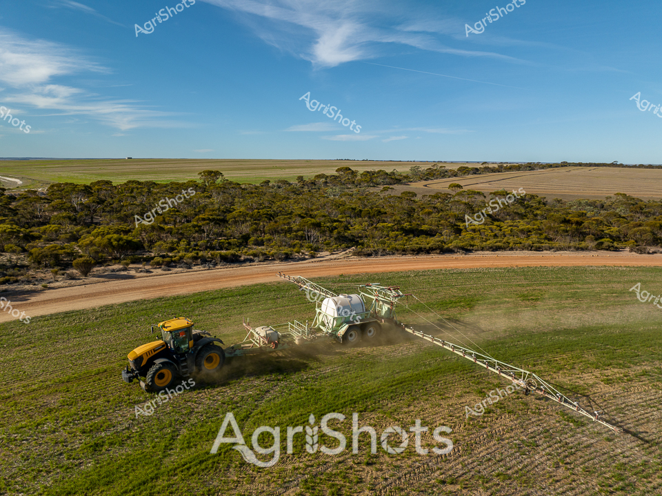 Sweeping across the undulating farmlands of Western Australia, a vibrant yellow JCB tractor pulls a massive spray rig through a verdant green paddock. The powerful agricultural machine cuts a dramatic path between dense native bushland and carefully cultivated fields, kicking up a cloud of dust that highlights the precision of modern farming techniques. Gleaming white boom arms extend from the spray rig, dispersing agricultural treatments with mechanical efficiency under a brilliant azure sky adorned with wispy cirrus clouds. The landscape tells a story of careful land management - cultivated fields contrast against the rugged, textured scrubland of the Australian interior, revealing the delicate balance between agricultural development and preserved natural ecosystem. Soft golden light bathes the scene, suggesting early morning or late afternoon, when farmers typically conduct critical crop maintenance operations. This aerial perspective captures the scale and sophistication of contemporary agricultural practice in Western Australia, where technology and terrain merge in a dance of productivity and environmental stewardship.