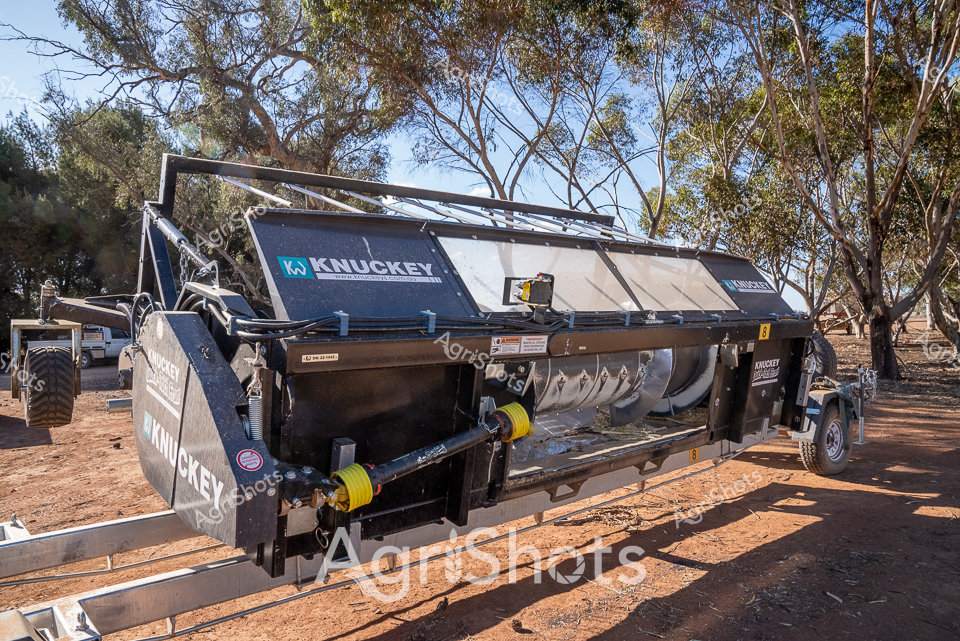 Gleaming under the dappled eucalyptus shade, a Knuckey harvest header front stands ready on dusty red earth, embodying the precision of modern Australian agricultural engineering. The metallic blue and grey machinery reflects the harsh sunlight, its complex mechanical design highlighting sophisticated grain harvesting technology. Robust yellow PTO (Power Take-Off) coupling points contrast against the dark machinery frame, suggesting industrial strength and immediate operational readiness. Positioned in what appears to be a typical dry Australian paddock—with sparse, weathered gum trees and packed ochre soil—this agricultural implement represents the cutting edge of harvesting equipment designed specifically for challenging rural conditions. The Knuckey branding prominently displayed signals a quintessential Australian agricultural manufacturing heritage, showcasing equipment engineered to withstand extreme harvesting environments. The header front's sleek, angular design speaks to both functional efficiency and technological innovation in contemporary grain production.