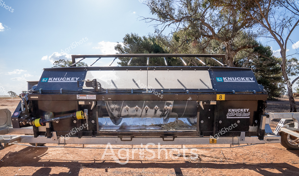 Stationed on the red-earthed plains of rural New South Wales, a Knuckey Quick-Tech Front harvesting header gleams under the bright Australian sunlight. The robust agricultural machinery stands resolute against a backdrop of weathered eucalyptus trees, casting angular shadows across the dusty ground. Branded prominently in navy and white, the header's industrial design speaks to precision agricultural engineering, with its substantial metal frame and intricate internal mechanisms designed for efficient crop processing. The number '8' marked distinctly on its side suggests this is part of a larger fleet of harvesting equipment, ready to tackle the challenging landscape of the Australian agricultural sector. Metallic surfaces reflect the intense midday light, creating a striking contrast against the muted earth tones of the surrounding landscape. This powerful piece of equipment represents the technological innovation driving modern farming practices in one of the world's most demanding agricultural environments.