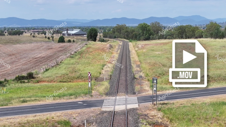 A rural railway crossing winds through the Liverpool Plains region of New South Wales, with verdant fields, distant mountain ranges, and a paved road intersecting the railroad tracks. The landscape showcases a classic Australian countryside scene, with gum trees, fenced paddocks, and a mix of agricultural and natural terrain under a soft, cloudy sky during what appears to be a warm summer or autumn day.