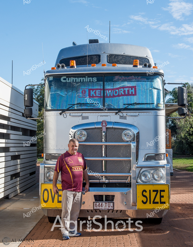 A seasoned Queensland truckie stands proudly beside his gleaming Kenworth prime mover, embodying the spirit of long-haul transport in rural Australia. The immaculate white and blue truck, powered by a Cummins engine, dominates the foreground, adorned with oversized yellow "OVER SIZE" warning signs that signal the vehicle's specialized freight capabilities. Wearing a maroon rugby jersey and casual pants, the driver exudes a sense of quiet confidence against the crisp blue sky and manicured surroundings. The truck's detailed chrome grille and pristine paintwork reflect meticulous professional pride, while the robust design speaks to the demanding conditions of agricultural freight transportation across vast Australian landscapes. Amber safety lights crown the cab, hinting at the vehicle's constant vigilance on remote highways. This portrait captures the intersection of industrial precision and personal commitment that drives Australia's critical agricultural supply chain, highlighting the unsung heroes who keep rural economies moving.