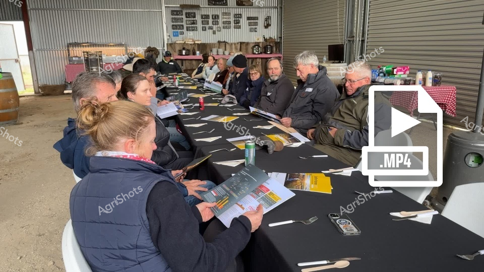 A group of agricultural professionals gather for a training seminar in a warehouse-like setting, with women and men seated around a large table reviewing safety documents and materials. The participants, dressed in warm clothing, appear focused on the discussion, with papers, notebooks, and beverages spread out before them, suggesting an informative workshop focused on agricultural safety and industry practices in New South Wales.