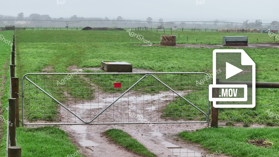 A weathered steel gate marks the entrance to a misty Tasmanian dairy pasture on a crisp winter morning. Muddy wheel tracks wind through lush green grass, revealing the well-worn path between paddocks and milking sheds. Heavy fence lines define the pasture's boundaries, with distant farm structures barely visible through the soft grey atmospheric haze. Utilitarian feed troughs and shelters are strategically placed across the undulating landscape, suggesting careful livestock management. The saturated ground and soft light communicate the damp, cool conditions typical of Tasmanian winter farming. A small red marker on the gate adds a subtle punctuation to the muted green and grey palette, hinting at systematic farm recordkeeping and agricultural precision.