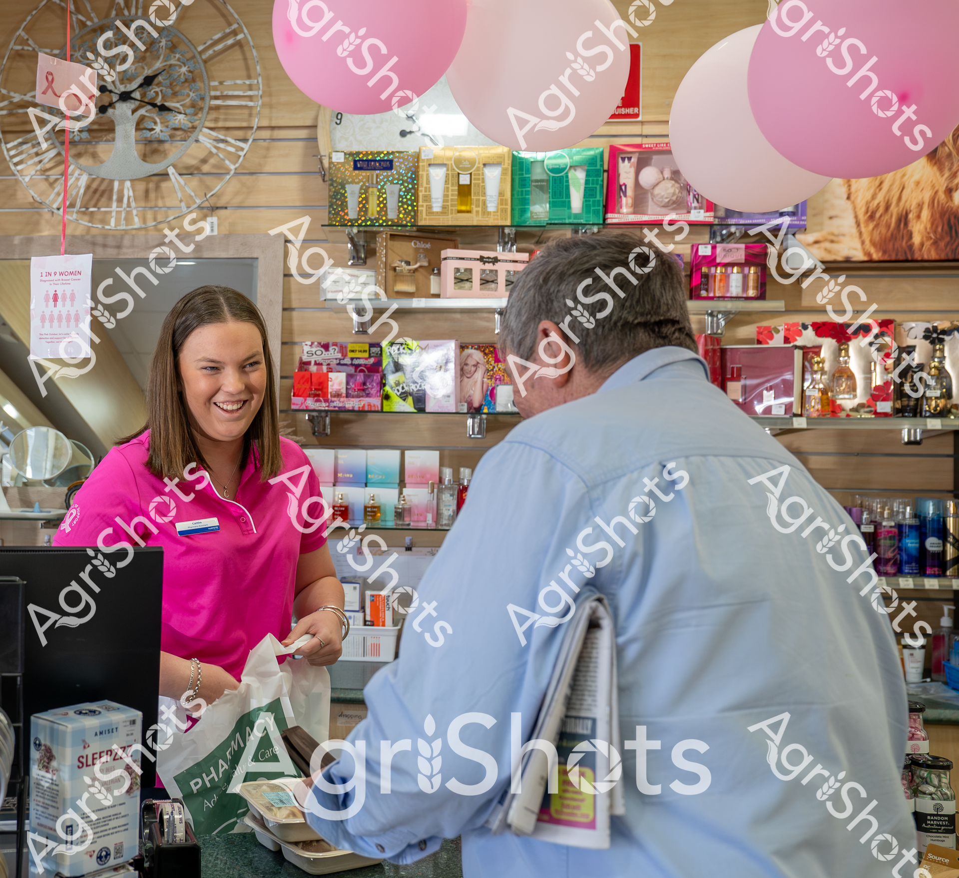 A friendly woman in a bright pink shirt stands behind a pharmacy counter, smiling while helping a customer in a blue shirt. The store is decorated with pink balloons and shelves stocked with various health and beauty products, creating a welcoming retail environment.