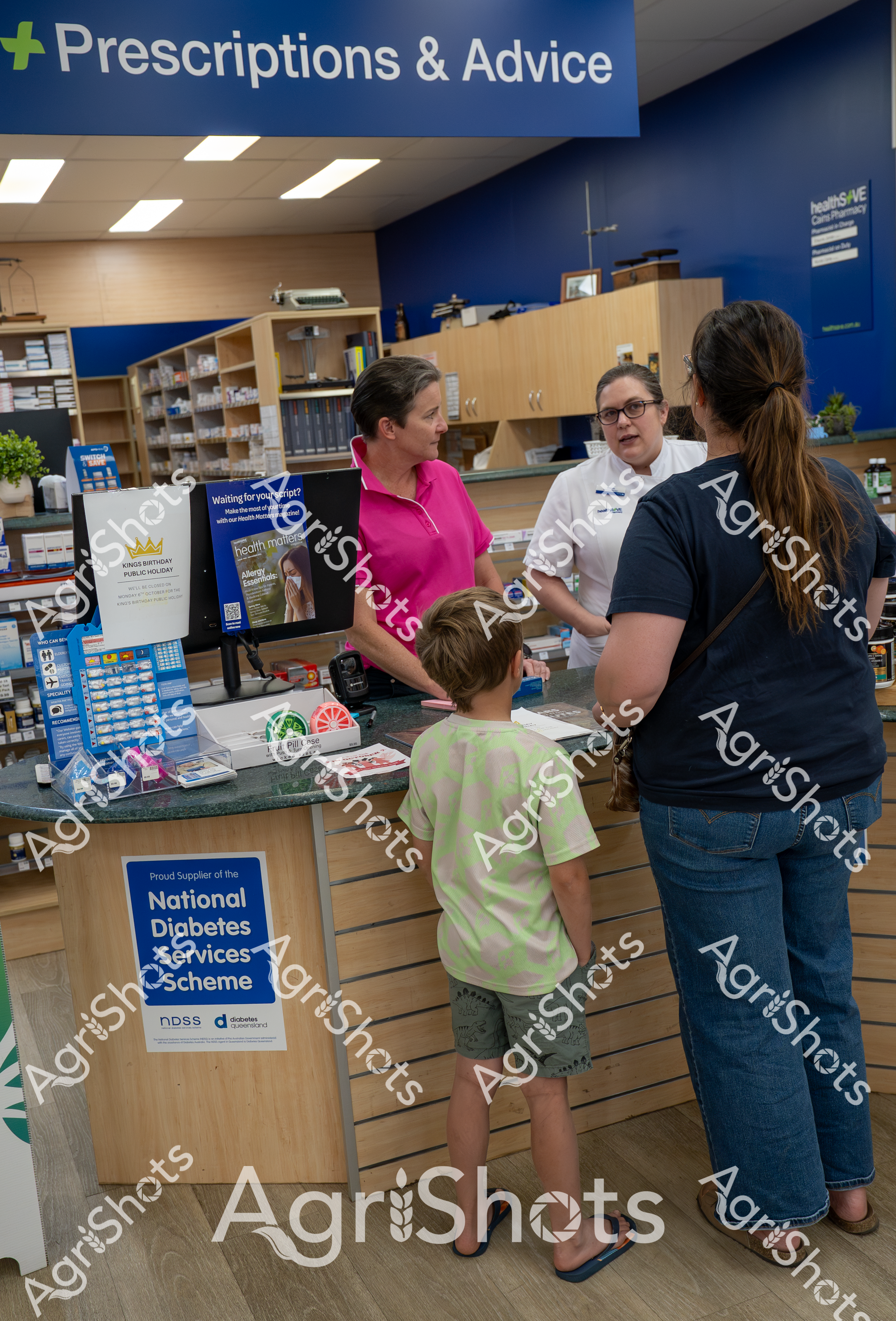 At a pharmacy's "Prescriptions & Advice" counter, a woman in a white coat speaks with a woman in a pink shirt and a child, discussing health services. The pharmacy displays a sign for the National Diabetes Services Scheme, indicating their commitment to supporting patients with diabetes care.