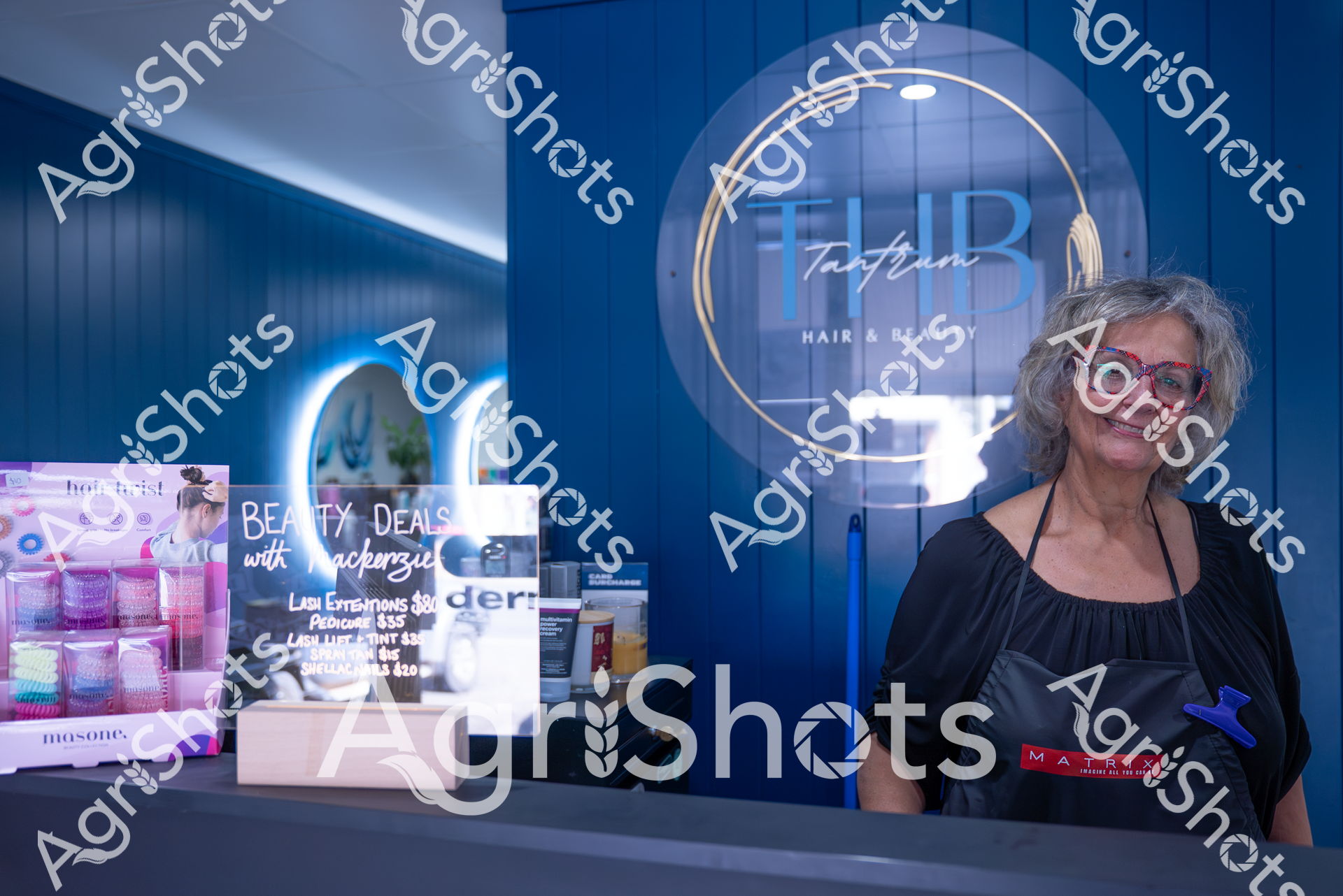 A mature woman stands in a hair salon with a warm smile, wearing glasses and a black top. The background showcases beauty product displays and the salon's branding, with blue walls and illuminated signage creating a professional atmosphere. She appears to be an employee or stylist, positioned near promotional materials for beauty treatments and extensions.