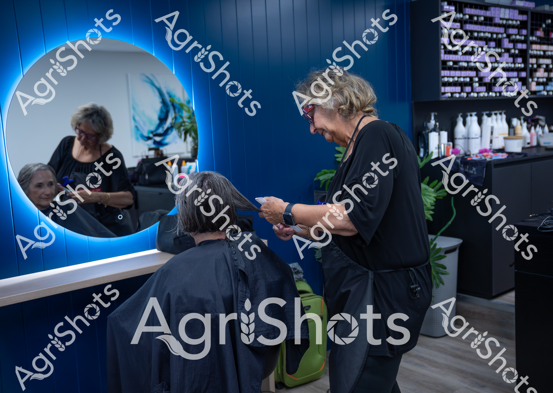 A seasoned hairstylist works on a client's hair in a modern, blue-themed salon. The stylist carefully trims and styles the client's hair while both are reflected in a large, illuminated circular mirror. The space is sleek and professional, with hair products and styling tools visible in the background.