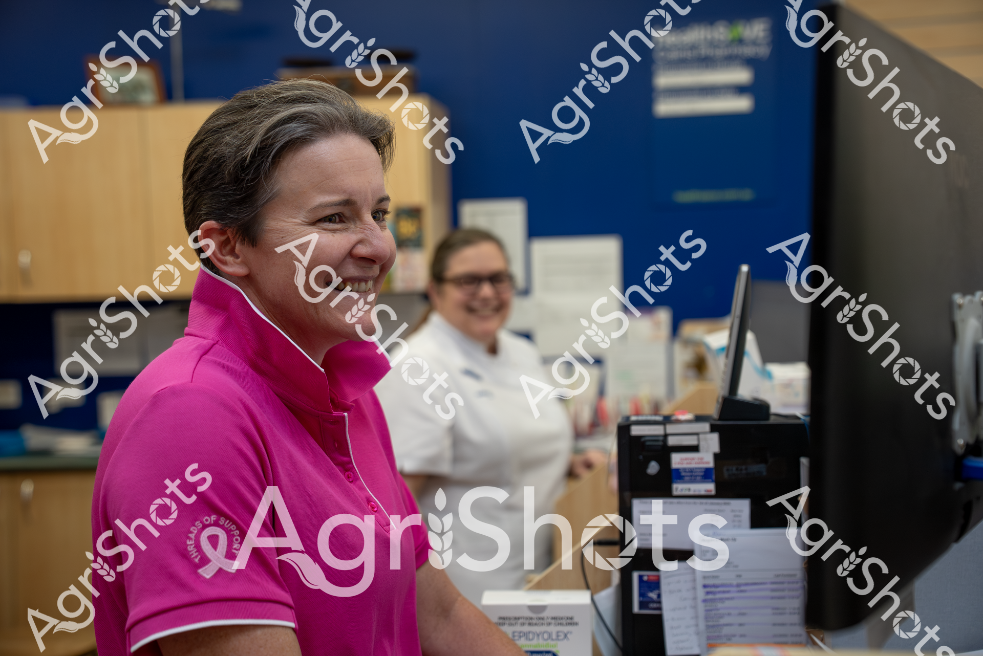 A woman in a bright pink top smiles warmly while working in an office environment. Behind her, another person in a white top can be seen working at a computer, with blue walls and office furniture in the background. The scene appears to be a professional workplace with a positive, friendly atmosphere.