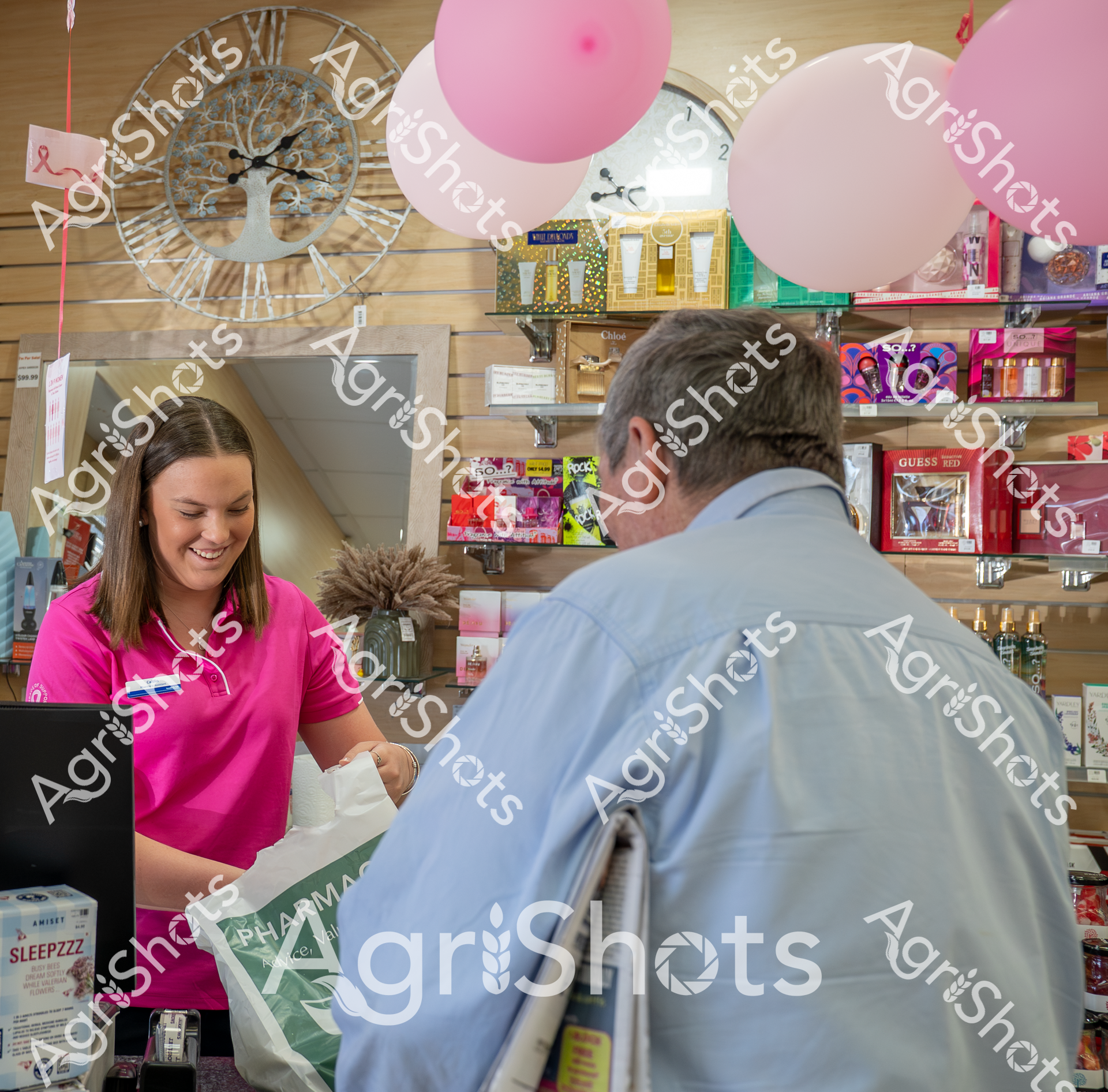A woman in a bright pink shirt assists a customer at a pharmacy or beauty store counter. Pink balloons and decorative shelves filled with beauty products create a cheerful retail environment. The interaction appears friendly and professional, with the employee smiling while helping the customer.