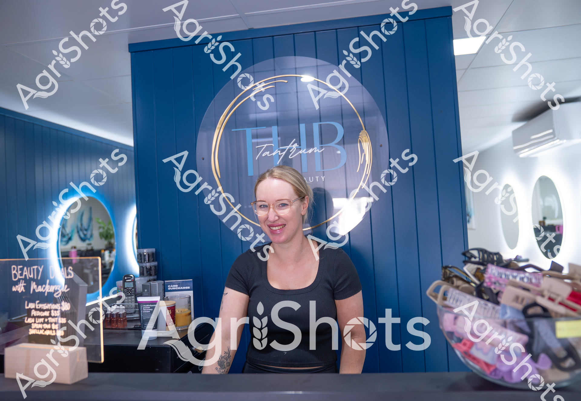 A smiling woman in a black t-shirt stands behind the counter of HB Tantrum Beauty, a blue-themed salon with various beauty products displayed. She wears glasses and appears welcoming, positioned in front of the salon's logo and signage. The interior looks modern and clean, with a professional and friendly atmosphere.
