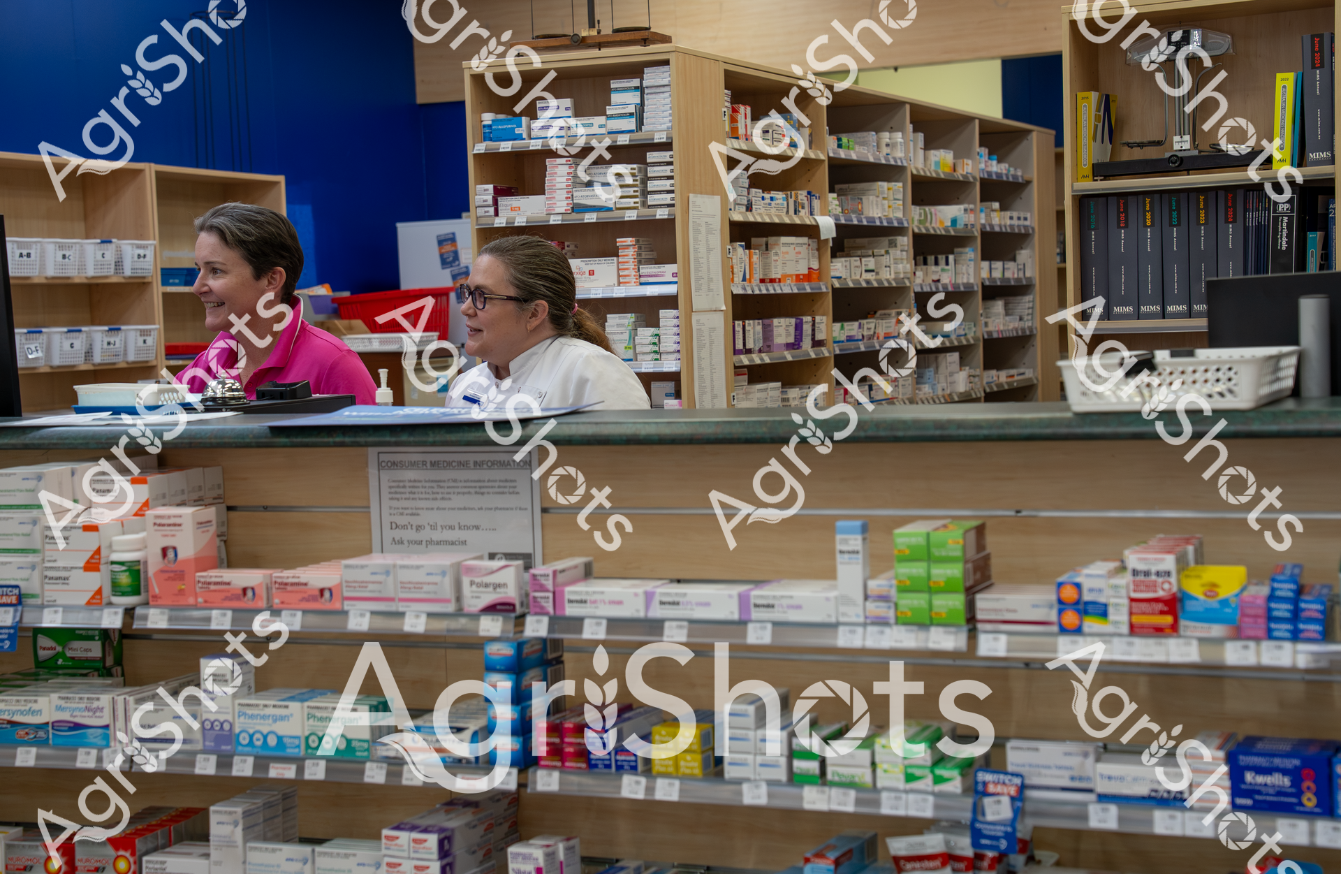 Two pharmacy workers are standing behind a counter filled with various medication boxes and shelves. They appear to be working in a well-organized pharmacy, with one woman in a pink top and another in a white coat, surrounded by neatly arranged pharmaceutical products and medical information displays.