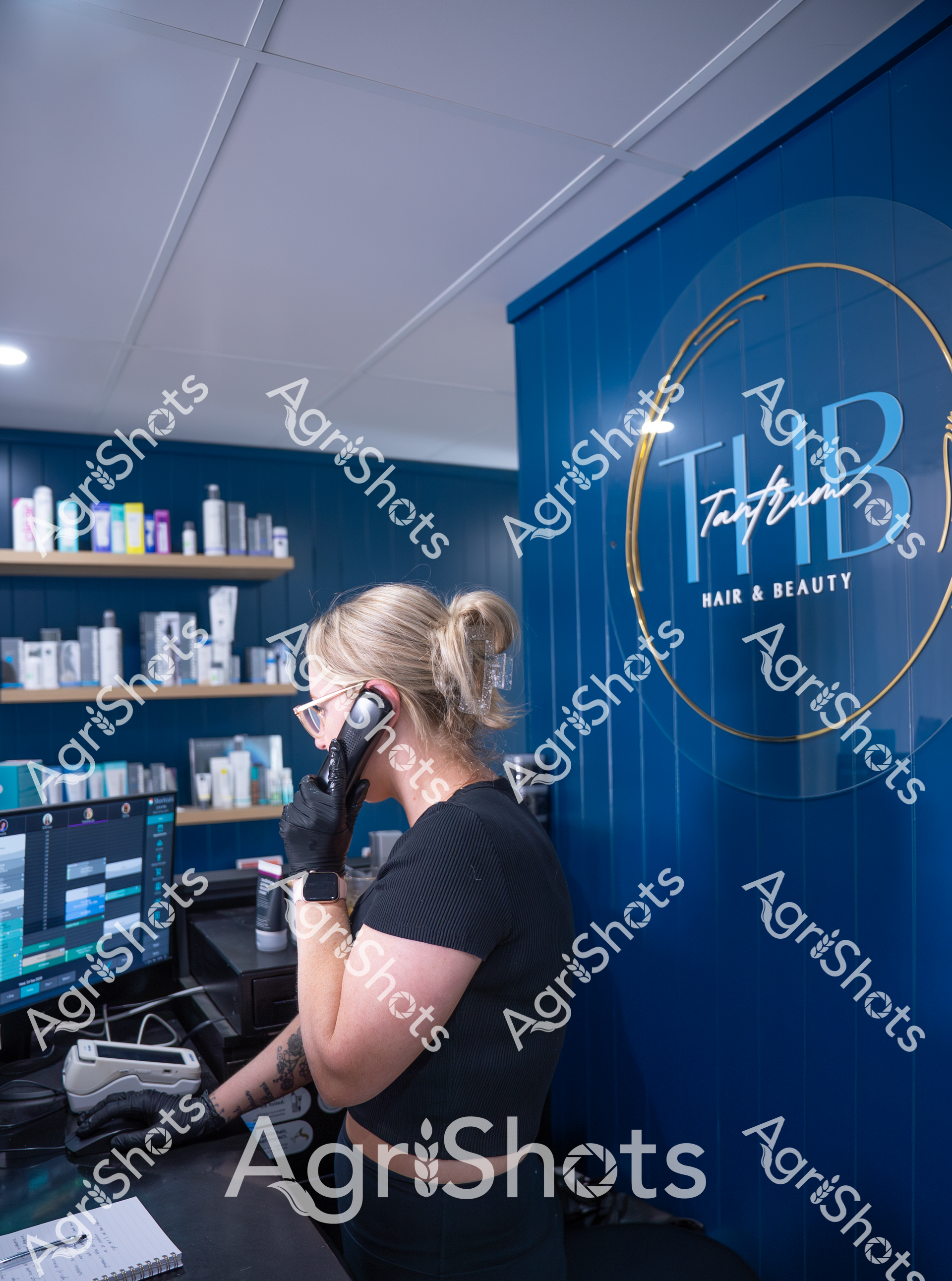 A woman with her hair in a bun wearing black gloves stands at a computer in a hair and beauty salon. She is looking at a screen and appears to be working, surrounded by hair products and a blue wall with a circular logo. The interior looks modern and professional, with shelves of hair care products in the background.
