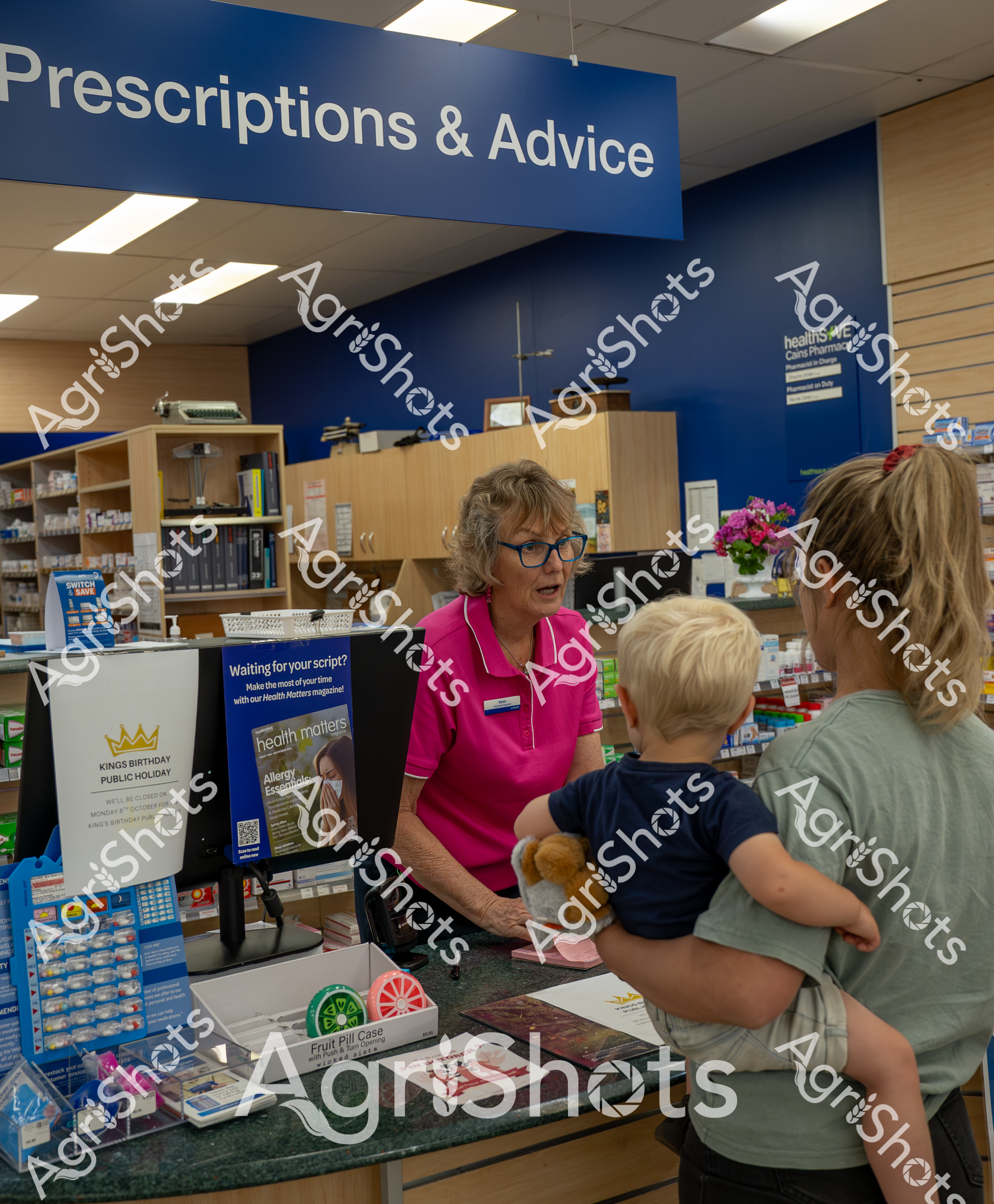 A woman in a bright pink shirt assists a customer at the Prescriptions & Advice counter of a pharmacy. A small child with blonde hair stands nearby, holding a stuffed animal, while another person is partially visible. The pharmacy setting includes shelves of medications, informational posters, and a clean, organized workspace.