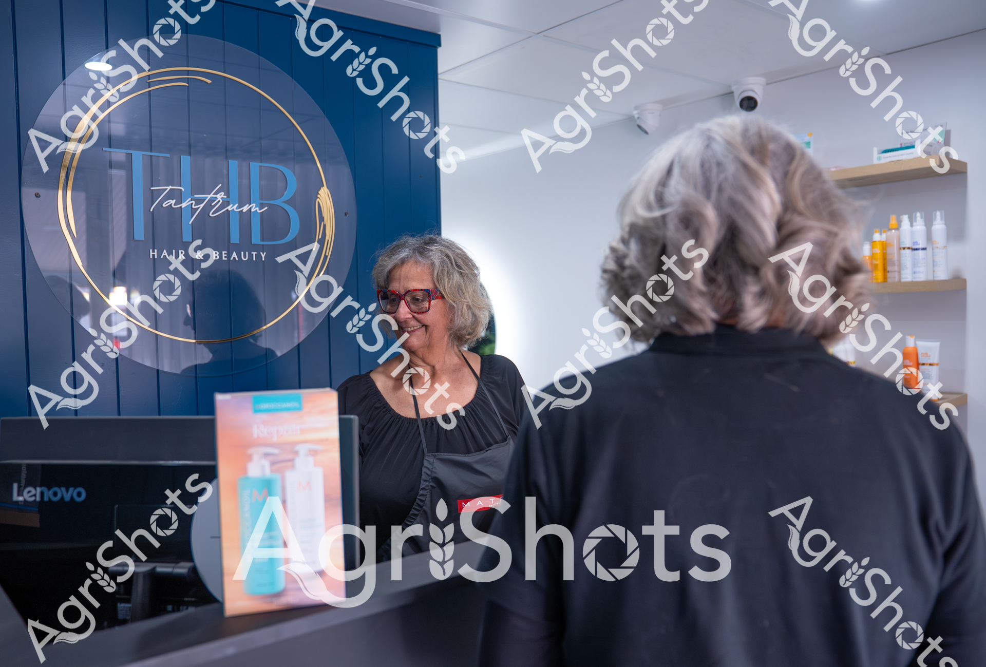 A smiling woman with gray hair and red glasses stands behind the counter at a hair and beauty salon called THB (Tantrum Hair Beauty). She appears to be interacting with another person whose back is facing the camera, with product displays and salon equipment visible in the background.