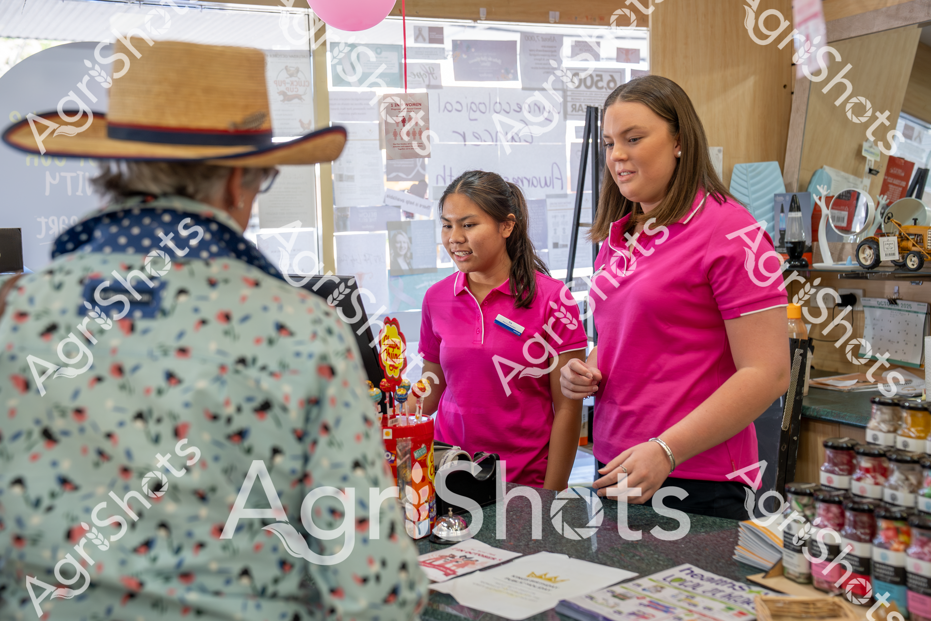 Two women in bright pink polo shirts stand at a counter, assisting a customer with colorful displays and products around them. They appear to be working in a retail or service environment, with a friendly and helpful demeanor. A pink balloon adds a cheerful touch to the scene.