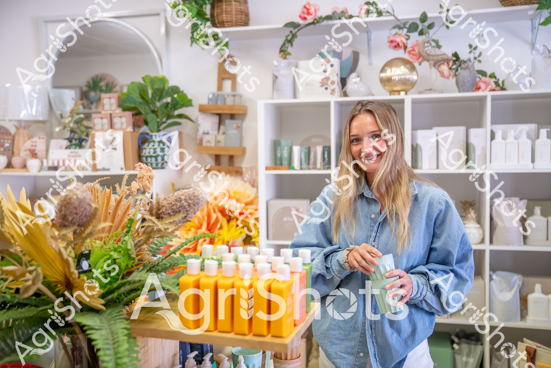 A woman in a denim shirt stands in a beautifully styled retail space filled with botanical products, surrounded by yellow bottles, dried flowers, and elegant shelving with decorative items. She smiles warmly while holding a mint green product, showcasing a welcoming atmosphere in what appears to be a boutique or wellness store with a natural, organic aesthetic.