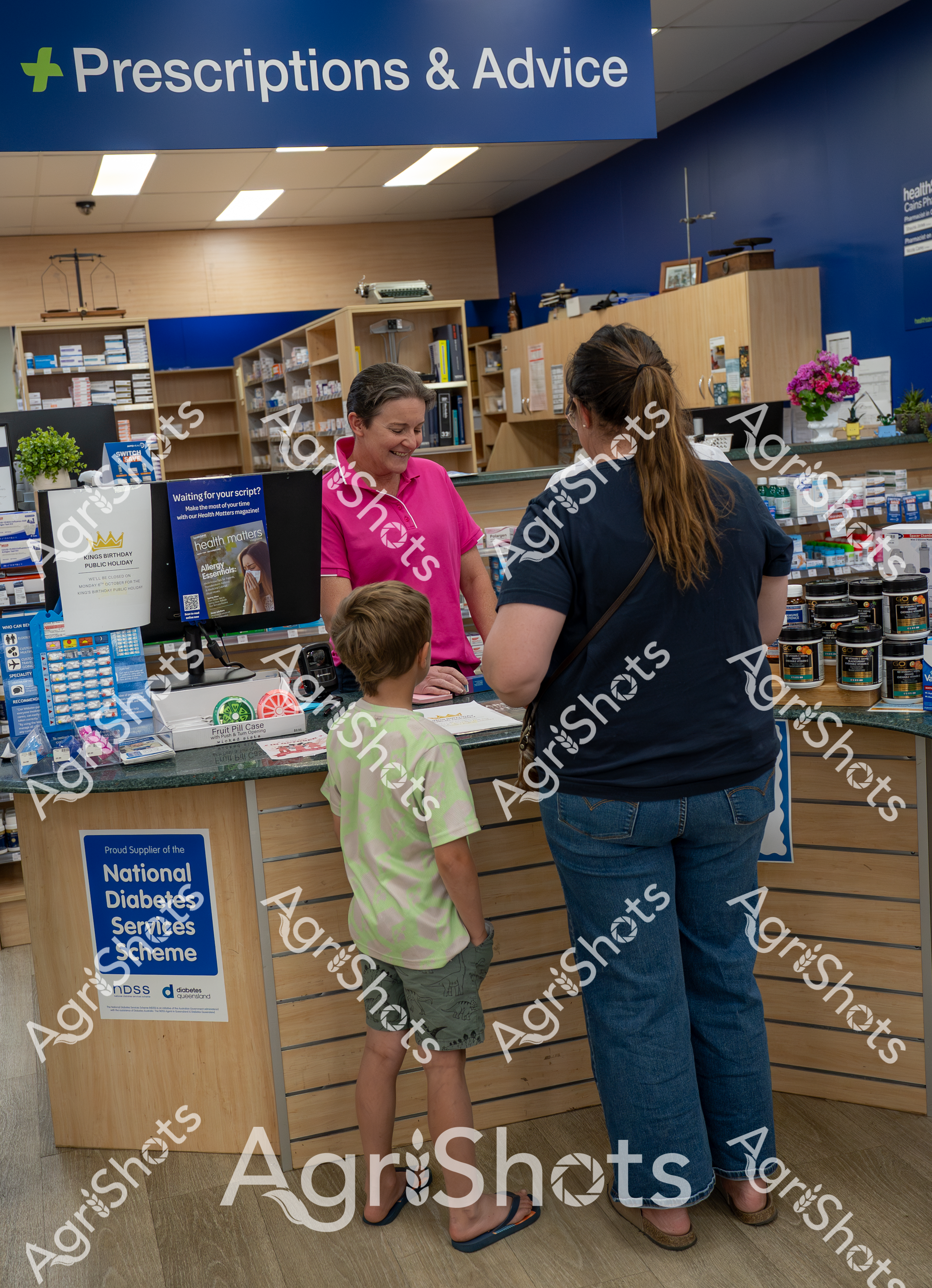 In a pharmacy's "Prescriptions & Advice" section, a woman in a bright pink shirt assists a customer with a child at the counter. Shelves of medications and health products line the walls, and a sign indicates the pharmacy is a proud supplier of the National Diabetes Services Scheme.