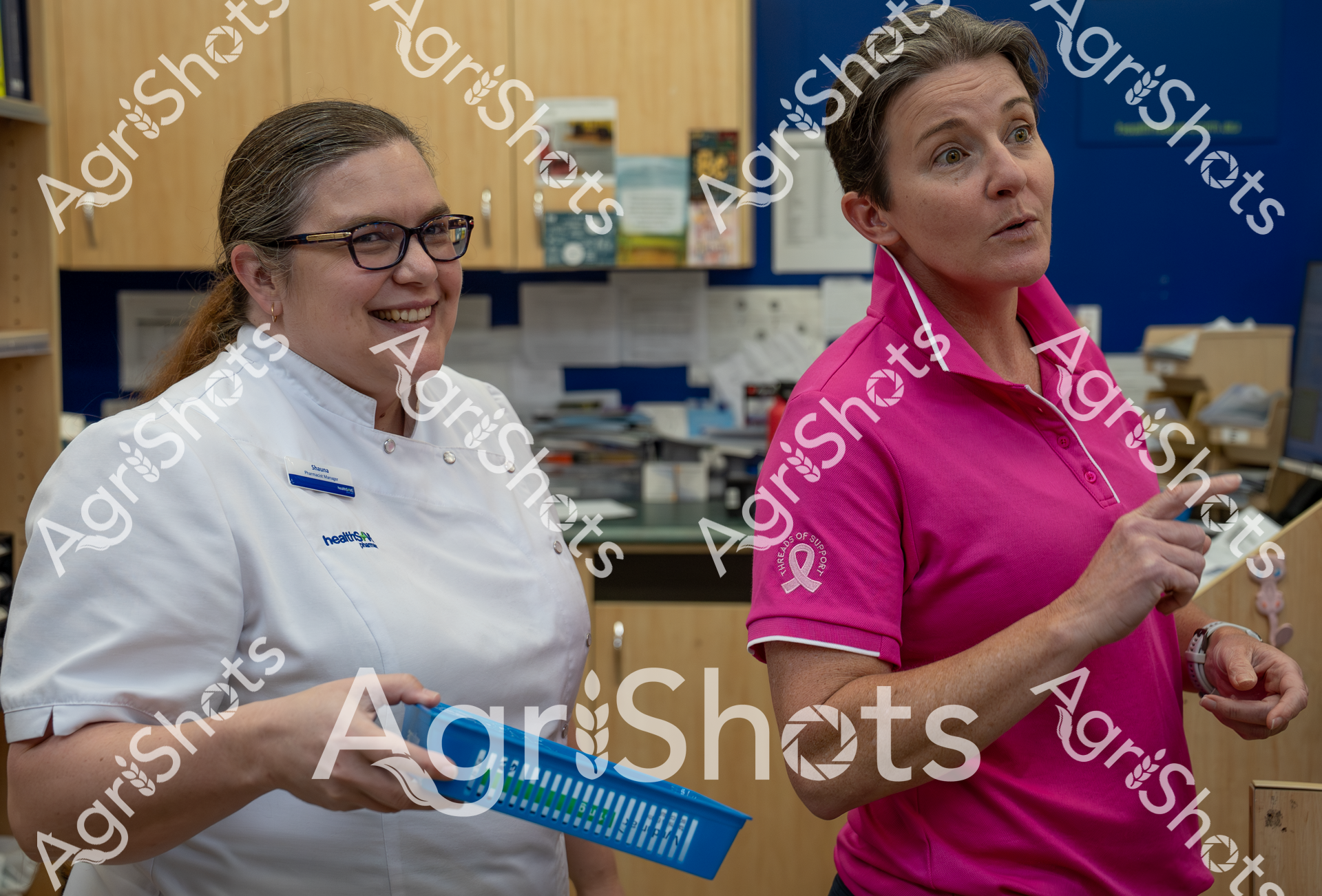 Two women in professional attire stand in an office or medical setting. One wears a white healthcare uniform and glasses, smiling, while the other is dressed in a bright pink shirt and appears more serious. They seem to be working together in a workplace environment.