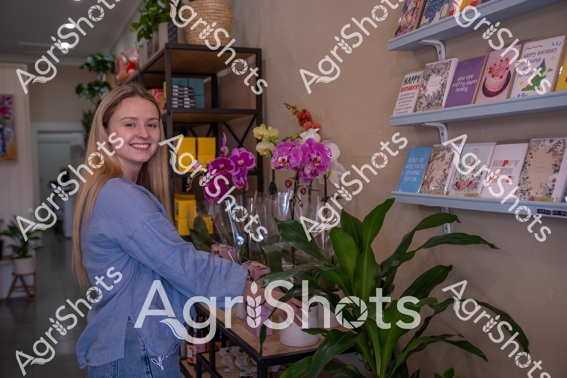 A smiling woman in a blue top stands in a flower shop, arranging vibrant purple and yellow orchids. The shop features shelves of greeting cards and decorative plants, creating a warm and inviting atmosphere of creativity and natural beauty.