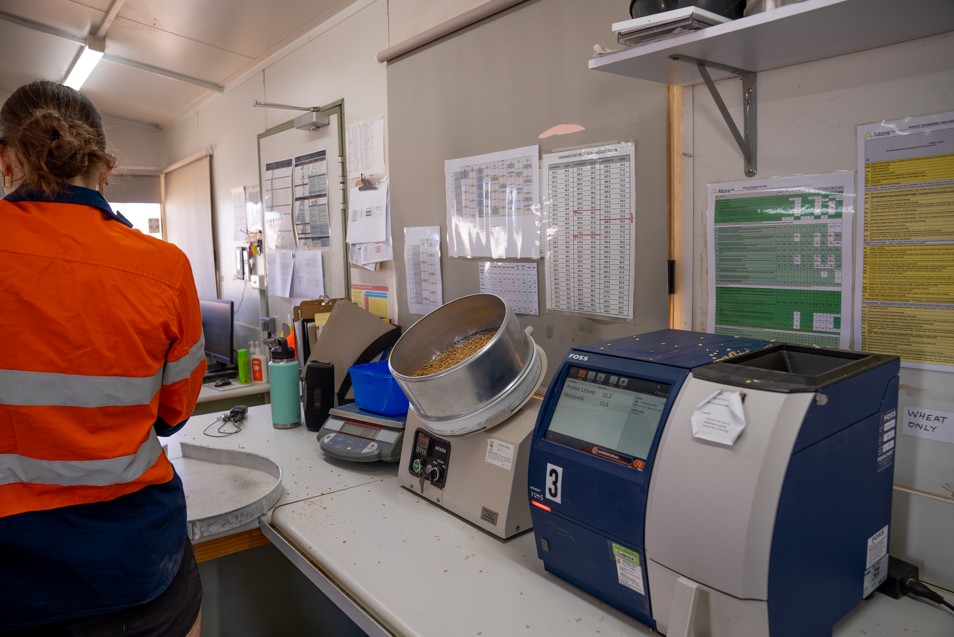 Agricultural Laboratory Quality Control Station: A worker in a high-visibility orange safety vest and reflective stripes stands at a grain testing workstation, surrounded by precision measurement equipment. A large metal sieve sits beside a digital scale, with golden wheat kernels ready for analysis, indicating a meticulous quality assessment process typical of modern grain production. The blue and white FOSS testing machine, marked with the number 3, represents cutting-edge technological instrumentation used to evaluate grain moisture, protein content, and other critical agricultural metrics. Crisp white walls are lined with charts, schedules, and operational protocols, reflecting the systematic and scientific approach of contemporary agricultural research and quality assurance. Multiple clipboards, calibration documents, and monitoring sheets underscore the rigorous documentation required in grain processing and export standards. The clean, organized workspace embodies the intersection of agricultural expertise and technical precision, highlighting Australia's sophisticated approach to agricultural science and grain industry standards.