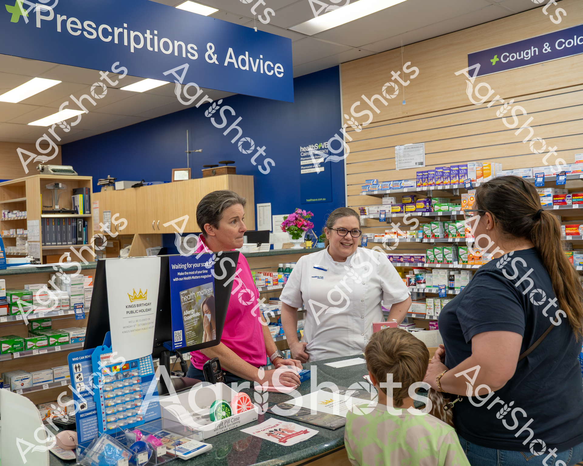A pharmacist in a white uniform stands behind the "Prescriptions & Advice" counter, assisting a woman and child. The pharmacy is well-stocked with medications and health products, with shelves of colorful boxes and informational displays in the background. The scene depicts a typical interaction at a local pharmacy, highlighting professional healthcare service.