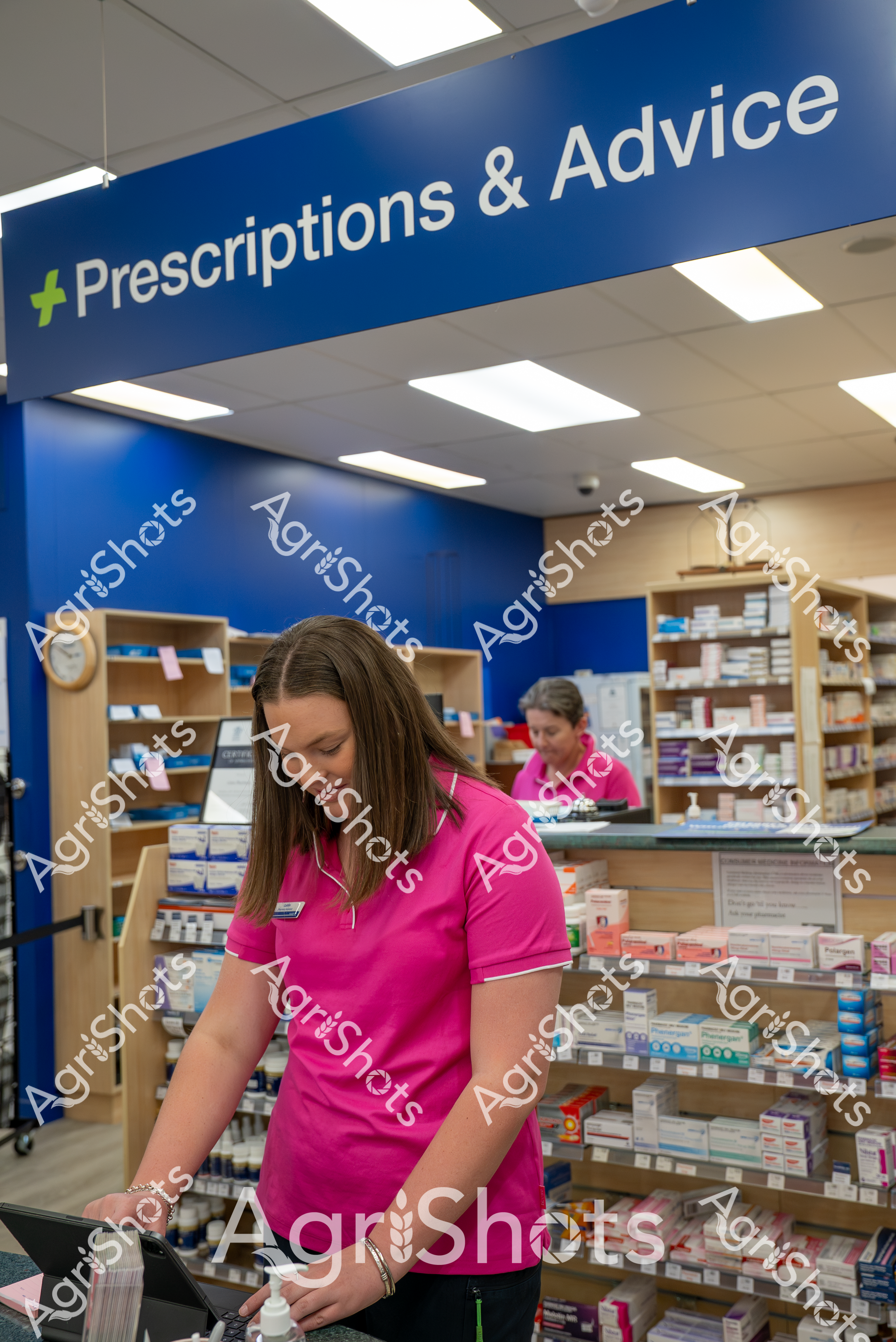 A pharmacist in a bright pink shirt works at the prescription counter of a pharmacy, surrounded by shelves of medication. The blue "Prescriptions & Advice" sign hangs overhead, indicating the dedicated area for patient medication services and pharmaceutical consultation.