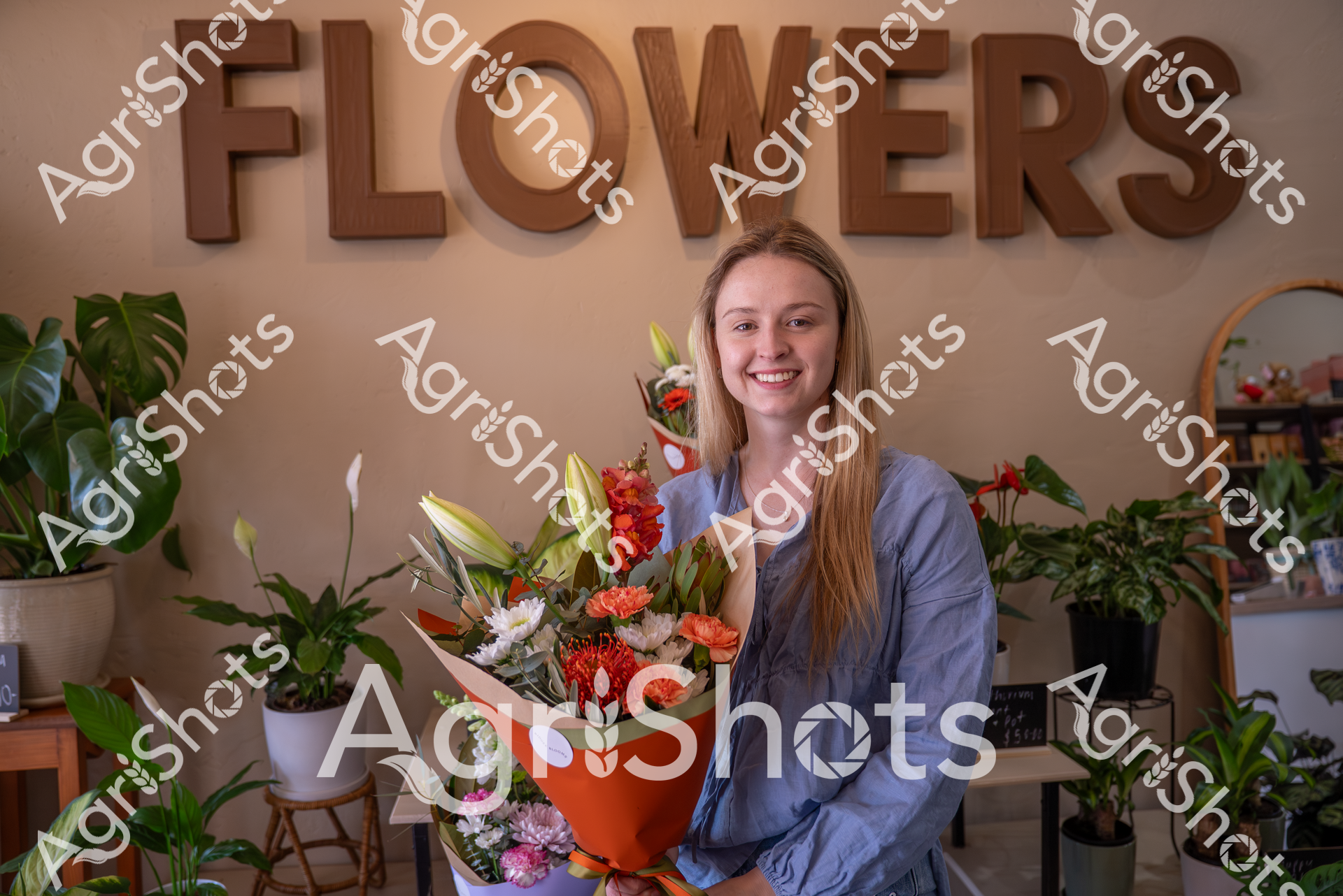 A smiling woman stands in a flower shop, holding a vibrant bouquet of colorful blooms. She wears a blue shirt and is surrounded by potted plants, with wooden "FLOWERS" letters displayed on the wall behind her, creating a warm and inviting atmosphere.