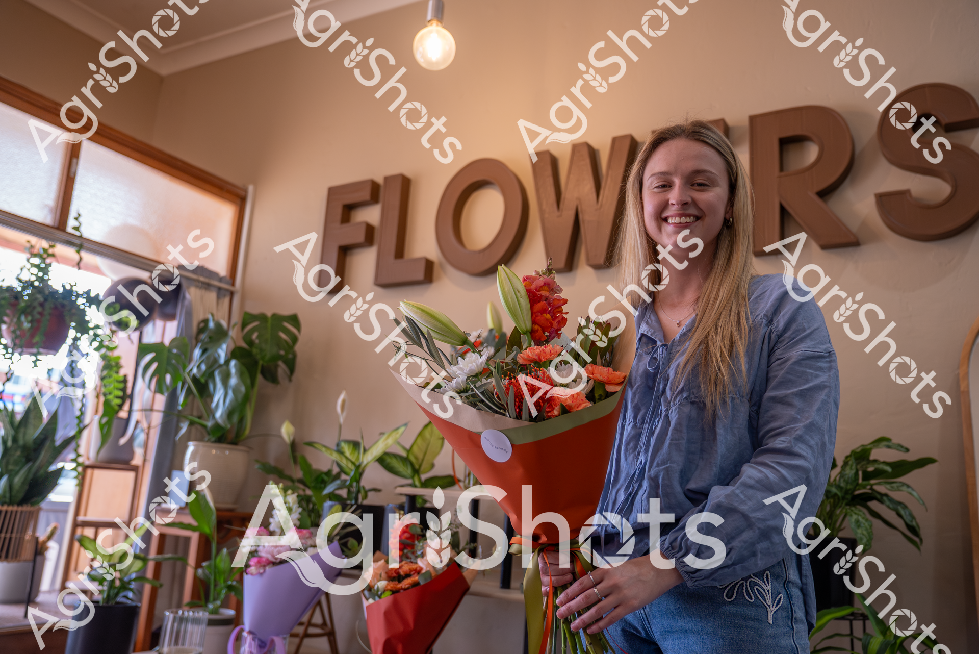 A smiling woman stands in a flower shop, holding a vibrant bouquet of red and white flowers. Surrounded by potted plants and beneath a large "FLOWERS" sign, she appears happy and content in her workplace, wearing a blue denim shirt.