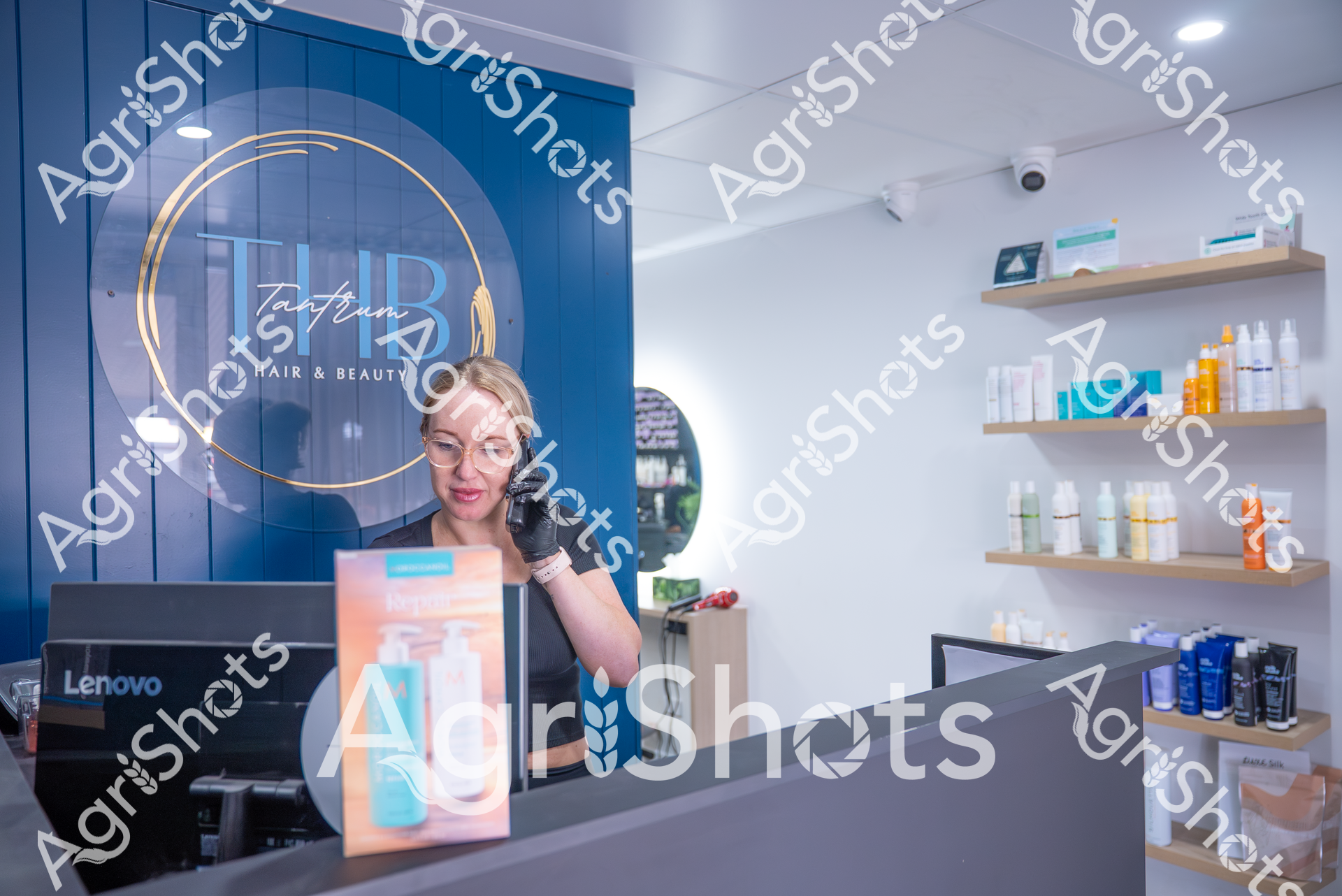 A woman wearing glasses and black gloves stands behind the counter of a hair and beauty salon called "THB Tantrum". Shelves in the background display various beauty and hair care products, and a Lenovo computer is visible on the counter. The salon has a modern, clean aesthetic with blue and white color tones.