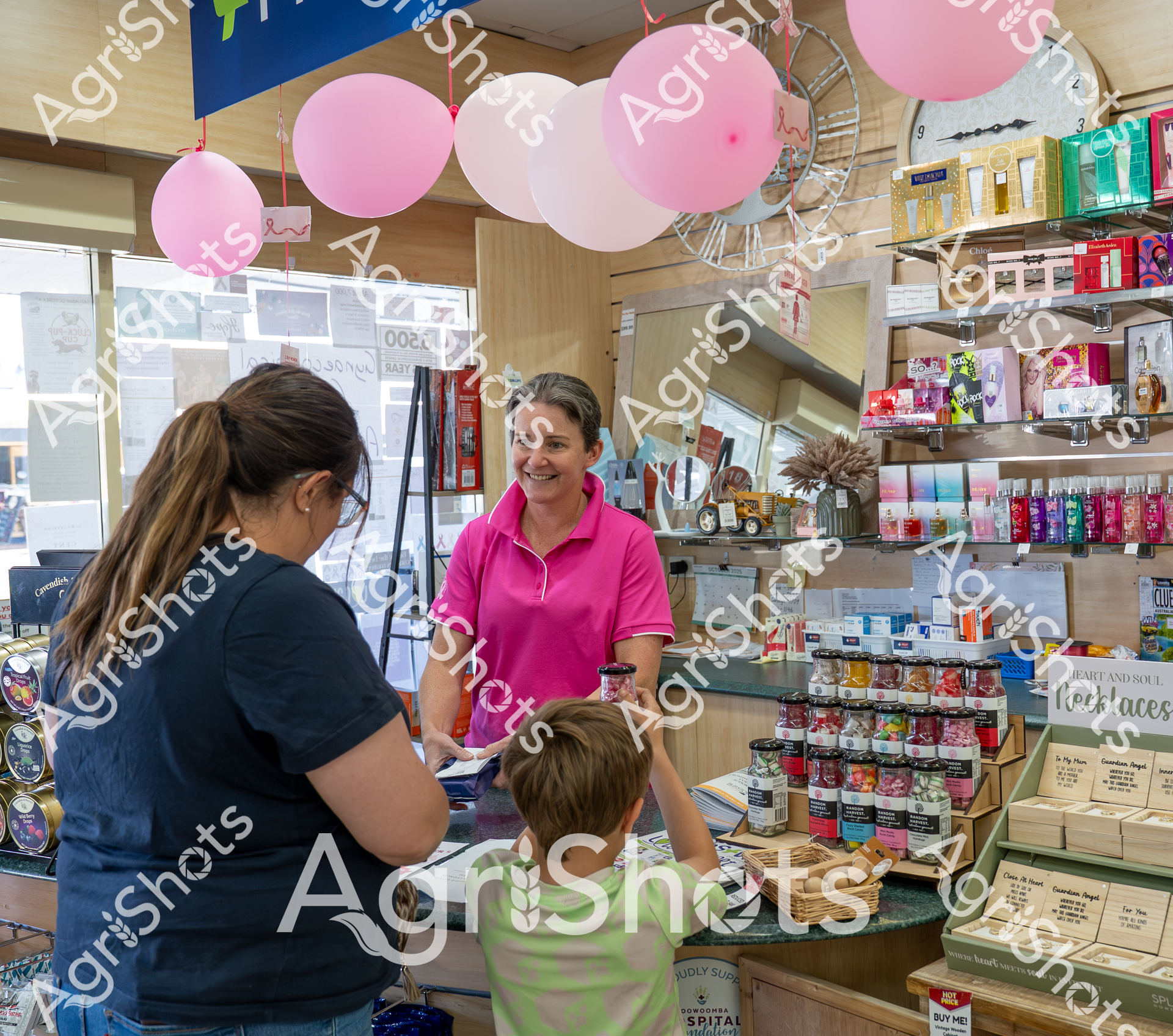 A cheerful woman in a bright pink polo shirt assists a customer at a retail counter, with pink balloons decorating the background. The store appears to be a gift shop or boutique, with shelves displaying various products like candles, jars, and packaged items. A child stands near the counter, adding to the warm, friendly atmosphere of the scene.