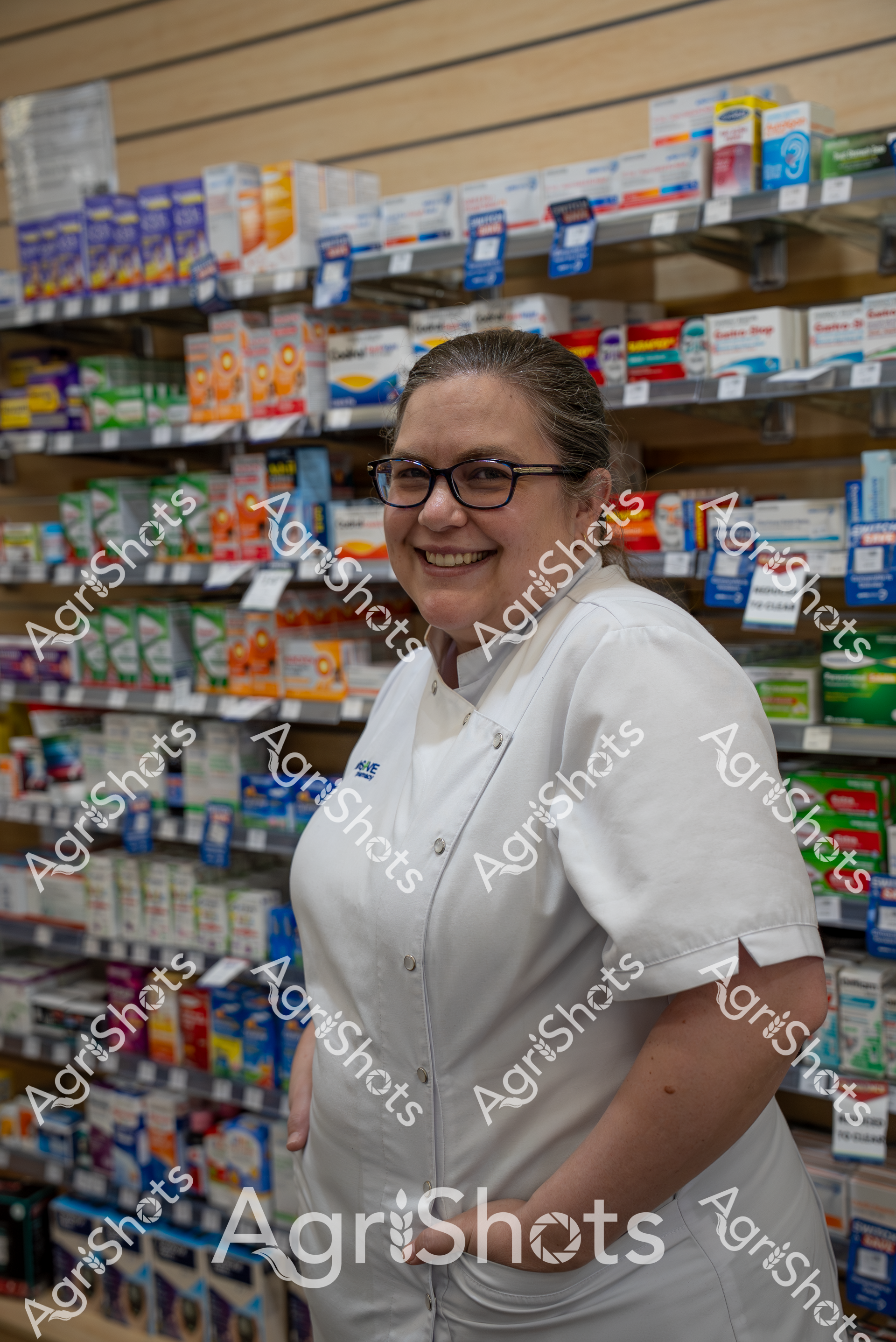 A smiling woman in a white pharmacy uniform stands in front of shelves filled with various medical and pharmaceutical products. She wears glasses and appears happy and confident in her workplace, surrounded by rows of medication boxes and healthcare supplies.