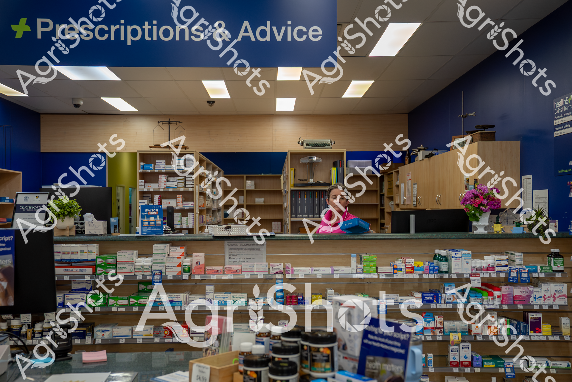 A well-stocked pharmacy counter with rows of colorful medication boxes and shelves. A woman in pink and blue attire stands behind the counter near a "Prescriptions & Advice" sign, surrounded by various pharmaceutical products and medical supplies.